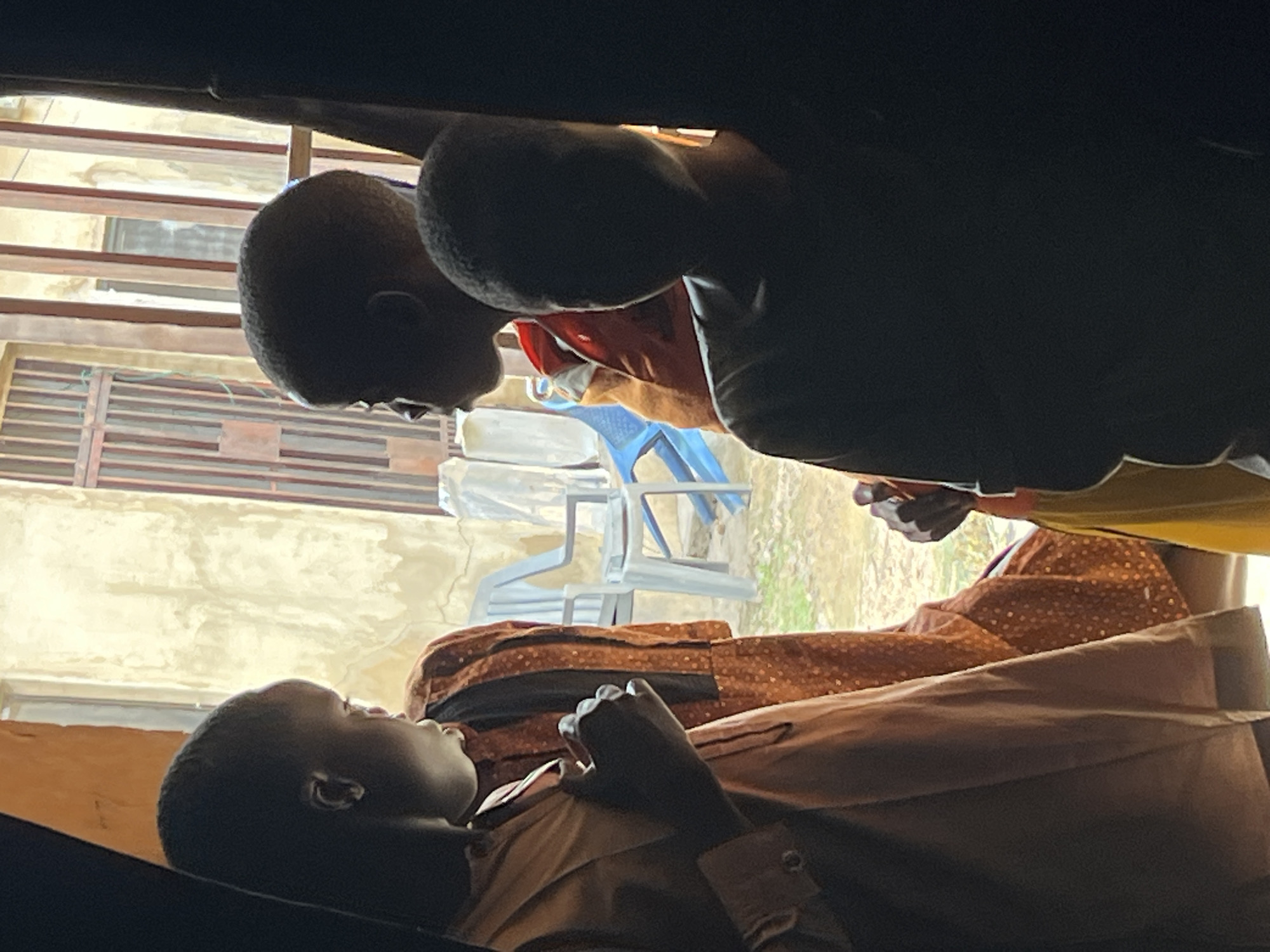 The image shows two young people, likely children, in an outdoor setting. One person is lying down, possibly on a cot or bed, wearing a light-colored shirt. Their head is tilted back, and their eyes are closed or looking upwards, suggesting they might be resting or sleeping. The other person is leaning over the first, their silhouette dark against a brighter background. We can see the back of their head and shoulders, and a hint of red clothing underneath.
The background reveals a textured, light-colored wall with horizontal bars or slats, possibly part of a fence or building. Two blue plastic chairs are visible further back, suggesting a casual, possibly communal area. The overall lighting appears to be natural daylight, and the atmosphere feels calm and perhaps a bit subdued. The image is oriented sideways, with the heads of the subjects appearing at the top and bottom of the frame. There is no visible text in the image.