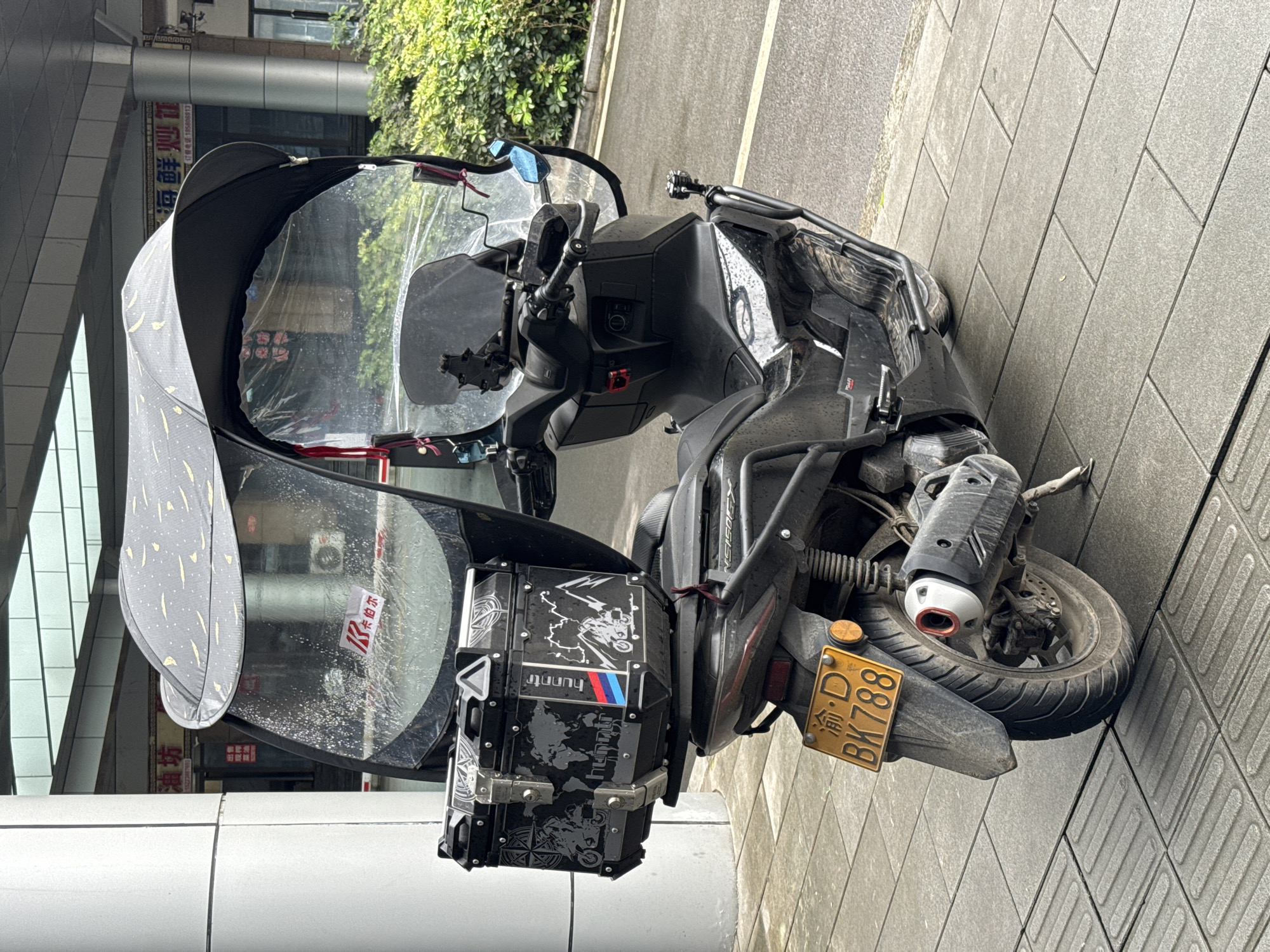 The image shows a black scooter parked on a gray-tiled sidewalk in Wulong District, China. The scooter has a large black canopy with silver and yellow patterns, and a clear windshield. A black storage box with various motorcycle-themed graphics and text is attached to the rear. The scooter's license plate is yellow and reads "渝·D BK788." Background details visible through the windshield include building facades and greenery. The scooter appears dusty or lightly soiled.
