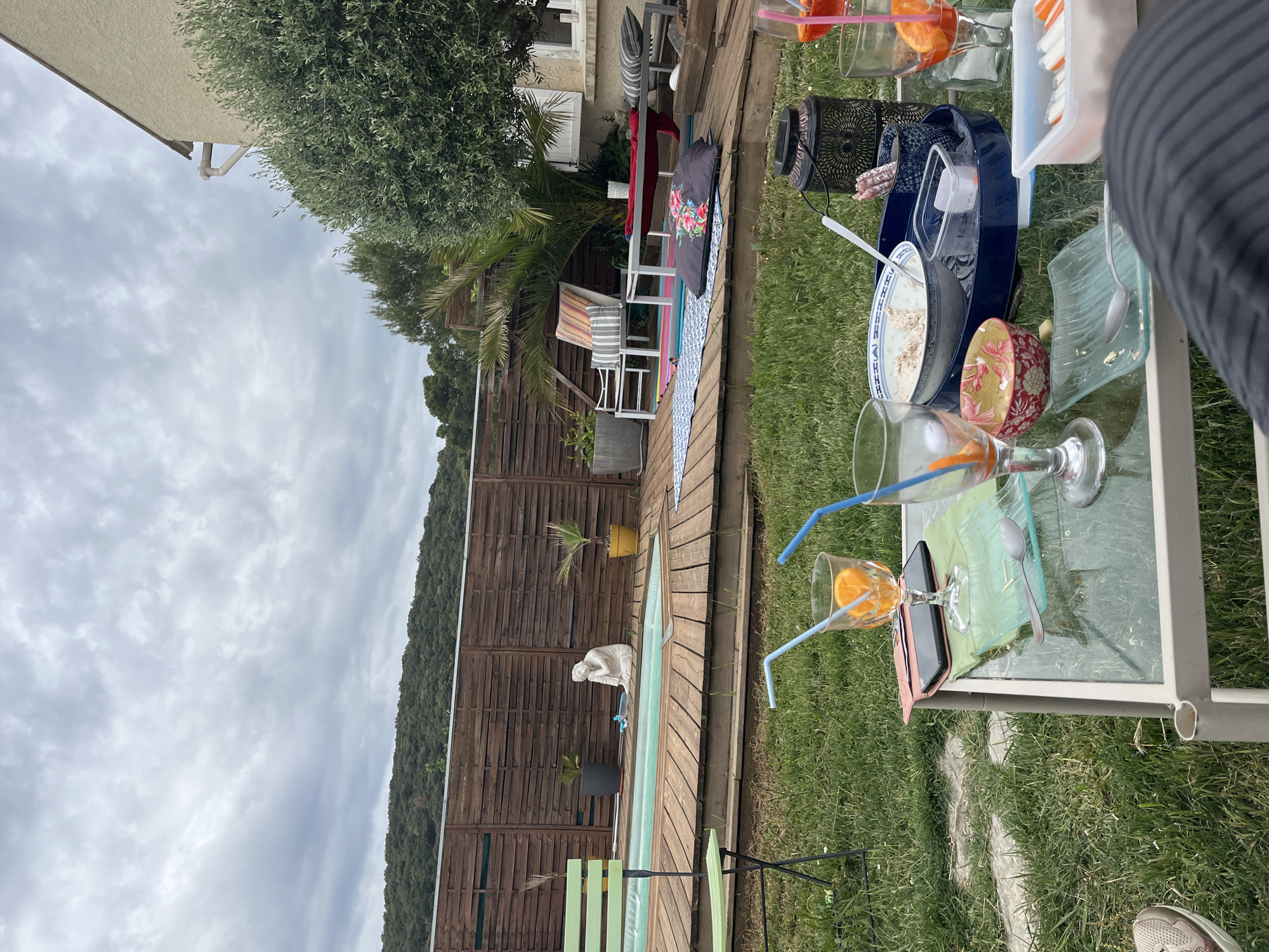 The image shows a backyard scene in Montarnaud, France. A rectangular wooden deck runs alongside a green lawn. On the deck, there's a narrow, light green swimming pool, a wooden fence, potted plants, and a white statue. Further into the backyard, there's a seating area with chairs and colorful blankets. A glass table is set up on the lawn, cluttered with used glasses containing orange slices and straws, plates with remnants of food, a bowl, and a cell phone. The sky is overcast. A person's arm and part of their leg are visible on the right side of the image.