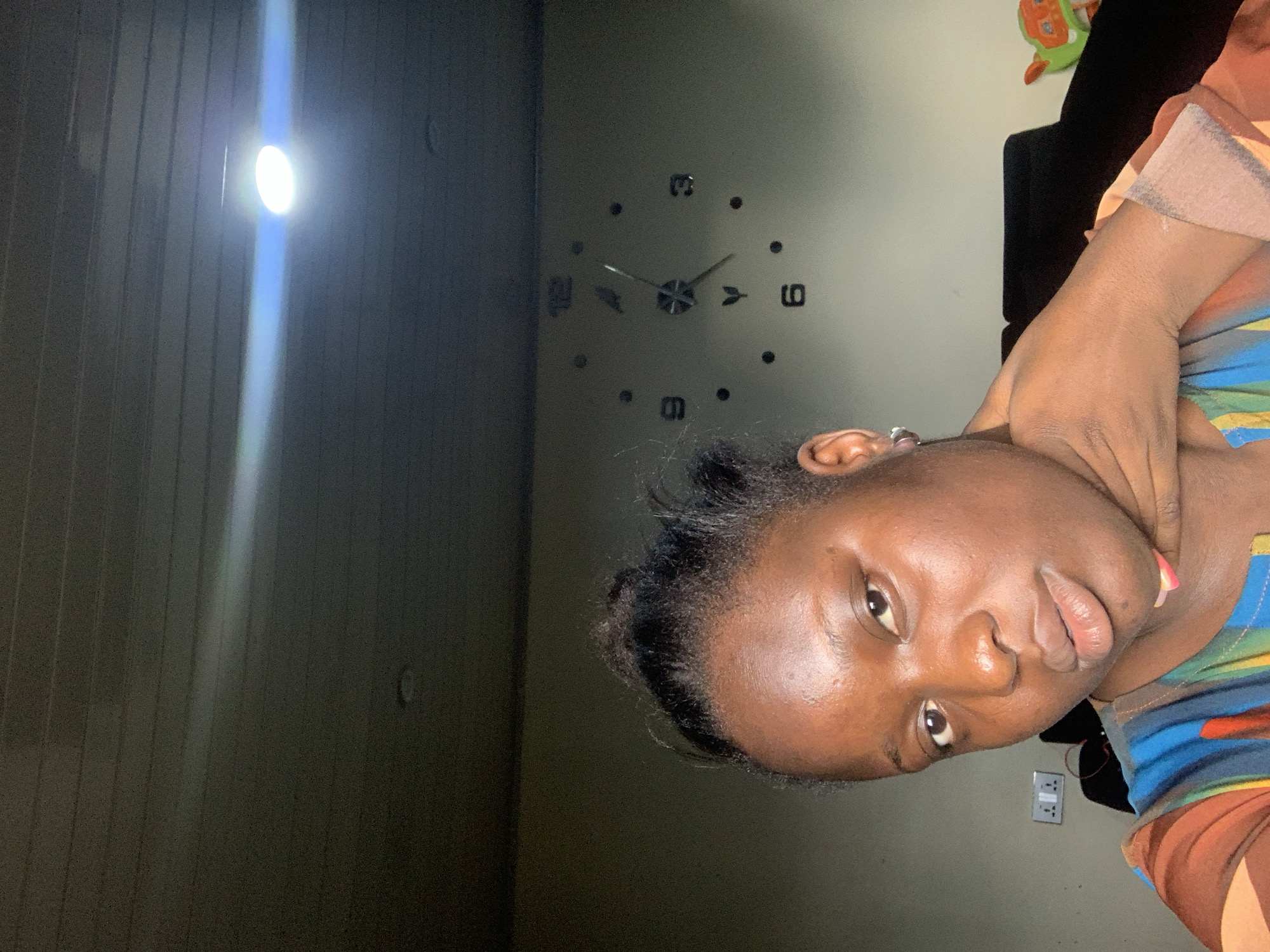 A young Black woman with her hair pulled up is shown from the chest up in a selfie. She is looking towards the camera with her right hand touching her neck and chin area. The woman is wearing a colorful shirt with stripes of brown, orange, blue, and yellow. In the background, there is a large wall clock with Roman numerals, a light switch, and the top of a dark couch. A light fixture is visible on the ceiling, emitting a bright glow.