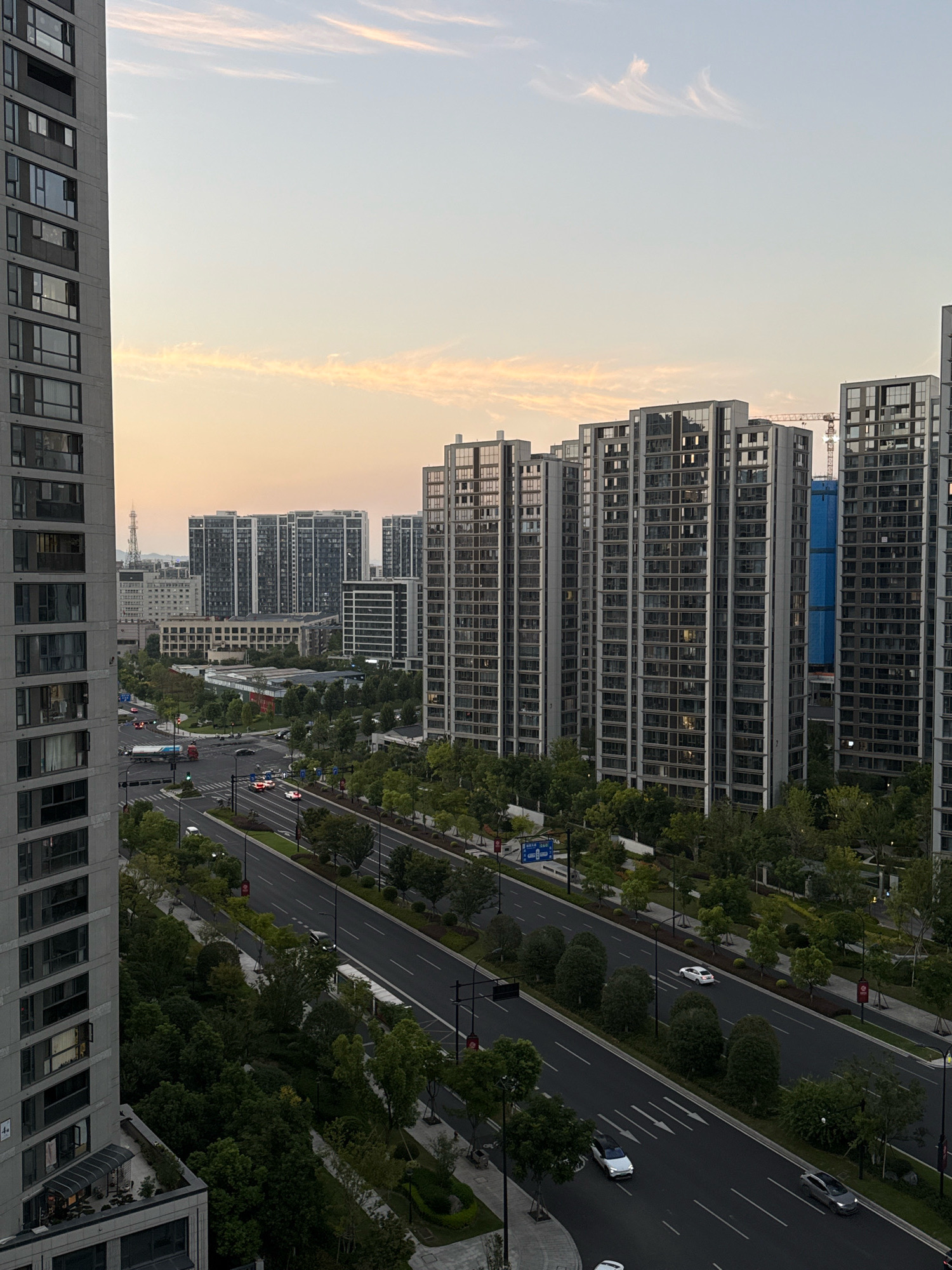 This is an aerial shot of a city street lined with tall, modern apartment buildings and lush greenery. The sky above is a soft gradient of pale blue and peach, with wispy clouds tinged with the warm glow of sunset. In the foreground, a tall, grey apartment building occupies the left side of the frame, its many windows reflecting the sky. Across the street, a wide, multi-lane road is visible, with a few cars and a truck moving along it. Trees and manicured landscaping line the road, creating a green buffer between the street and the rows of identical, high-rise apartment buildings that dominate the middle and right sections of the image. The buildings have a uniform design, featuring many glass windows and balconies. In the background, more buildings and a construction crane are visible, suggesting ongoing development in the city. The overall impression is one of a modern, well-developed urban environment bathed in the soft light of dusk.