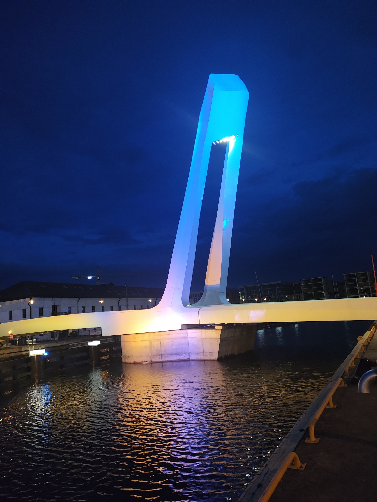 The image captures a nighttime scene of the Järve Bridge in Tallinn, Estonia, illuminated with vibrant blue and yellow lights. The bridge's distinctive modern architecture, with a tall, angular pylon glowing blue, is prominently featured against a deep blue, cloudy sky. The yellow pedestrian walkway of the bridge reflects warmly on the dark water below, creating a striking contrast with the cool blue illumination of the pylon. The water surface is textured with ripples, further enhancing the reflections. In the background, a low-rise building with a dark roof and illuminated windows is visible, suggesting an urban environment. A construction crane is also dimly visible in the distance. The foreground shows a dark walkway with yellow railings, indicating a viewpoint from the waterfront or pier. The overall impression is one of modern urban infrastructure bathed in dramatic artificial light.