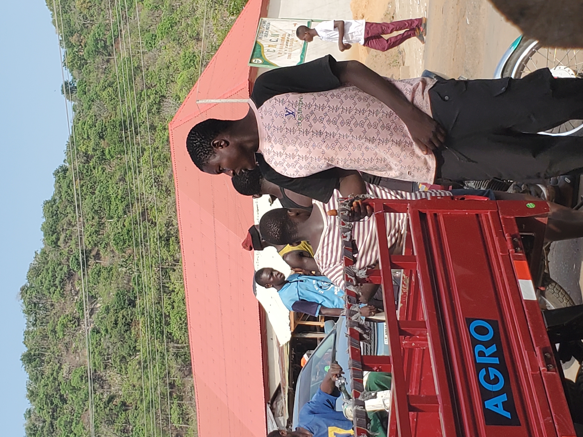The scene appears to be in Mararaba, Nigeria. A red cargo tricycle with "AGRO" prominently displayed on the side is occupied by several people. Some individuals are seated in the vehicle's cargo bed. The people are of African descent. One person is wearing a pink patterned shirt with black pants. A building with a red roof is nearby. A sign with text, partially obscured, is attached to the building. There is a hillside with dense green foliage in the background, and electrical wires run across the sky. Another person walks in the background. The general environment suggests a busy, outdoor setting along a street.