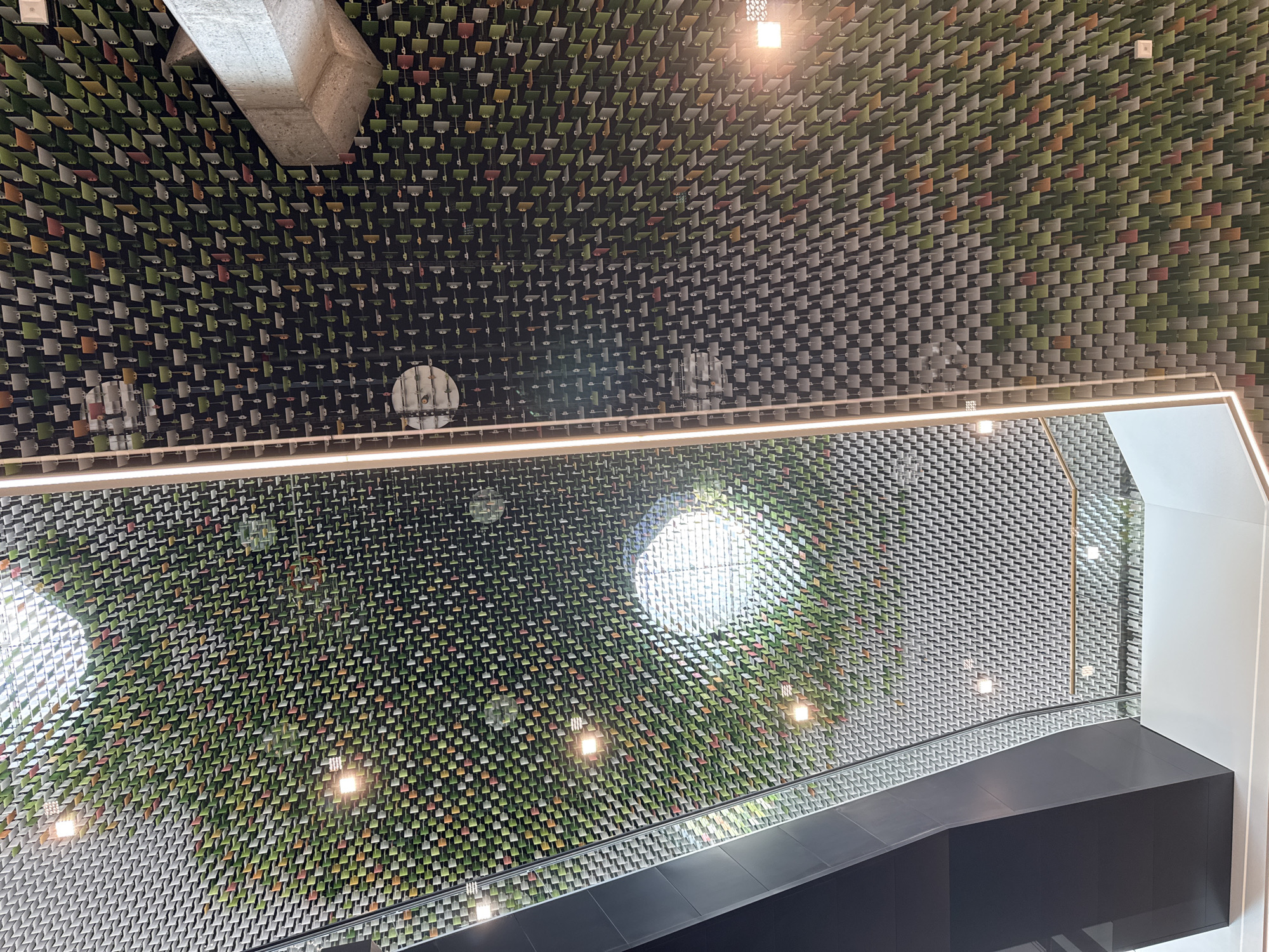 This image captures a ceiling structure in Morbio Inferiore, Switzerland, likely within a modern architectural space. The ceiling is a visually complex grid of small, rectangular elements, predominantly in shades of green, with interspersed accents of white, grey, and orange. These elements are arranged in a repeating pattern, creating a textured, almost mosaic-like effect.
Several circular light fixtures are visible, casting a warm glow onto the ceiling and the surrounding environment. A prominent concrete beam is also integrated into the ceiling structure, providing a stark contrast in material and form to the patterned elements.
To the left of the frame, a dark, angular architectural feature suggests an elevated walkway or staircase, partially obscured by a glass balustrade. This feature, along with the reflective surfaces of the ceiling, indicates an indoor setting, possibly a public building or commercial space. The overall impression is one of sophisticated design, utilizing repeating patterns and varied materials to create a unique aesthetic. There are no people visible in the image.