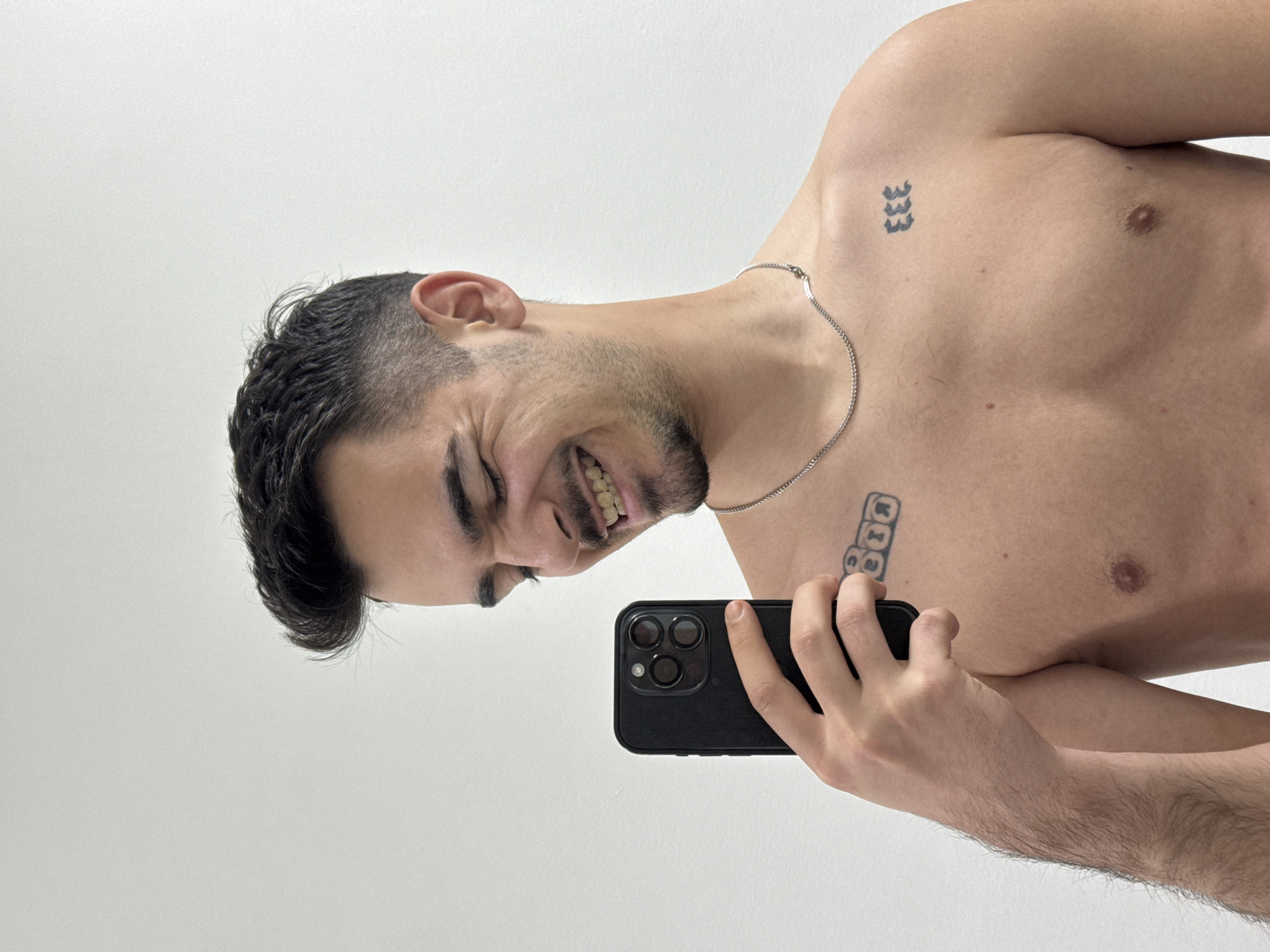 A young, shirtless man is depicted from the chest up, against a plain, light-colored background that appears to be an indoor wall. The original image is rotated 90 degrees counter-clockwise, showing the man's head facing towards the left side of the frame with his body extending to the right. He has short, dark hair and stubble on his face. His expression is one of strong amusement or joy, with his mouth open in a wide grin revealing his teeth, and his eyes squinted.
He is holding a black smartphone with multiple camera lenses in his right hand, positioned in front of his torso as if taking a selfie. A thin silver chain necklace is visible around his neck.
Notable details include two tattoos on his upper torso. On his left upper chest/shoulder, there is a tattoo of three stacked "3"s ("333"). On his right upper chest, there is another tattoo consisting of three letters or symbols enclosed in individual squares, though the specific letters are difficult to discern clearly due to the angle and rotation, potentially resembling "DDR" or similar. The lighting is even and soft. There are no specific visual cues within the image itself to indicate the provided location context of Sofia, Bulgaria.