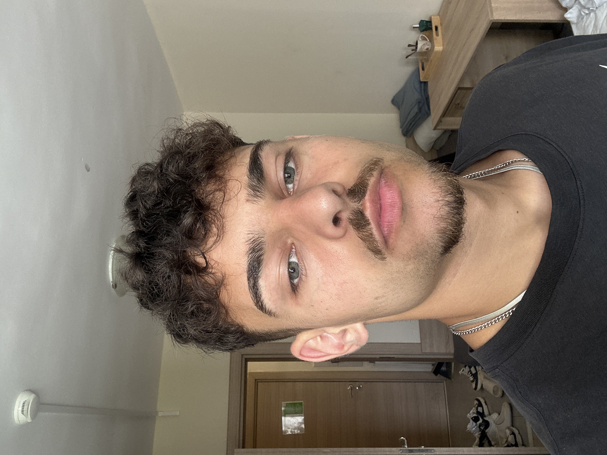 A close-up, tilted headshot of a light-skinned male with curly dark brown hair and a groomed mustache and beard. He has light-colored eyes. He is wearing a black t-shirt and two silver necklaces. The background includes a gray wall with a smoke detector, a wooden desk with items on it, a partially visible gray item, a door with a small window, and what appears to be a sink and a collection of sneakers or shoes. There is no information in the image to determine the location in Sofia, Bulgaria.