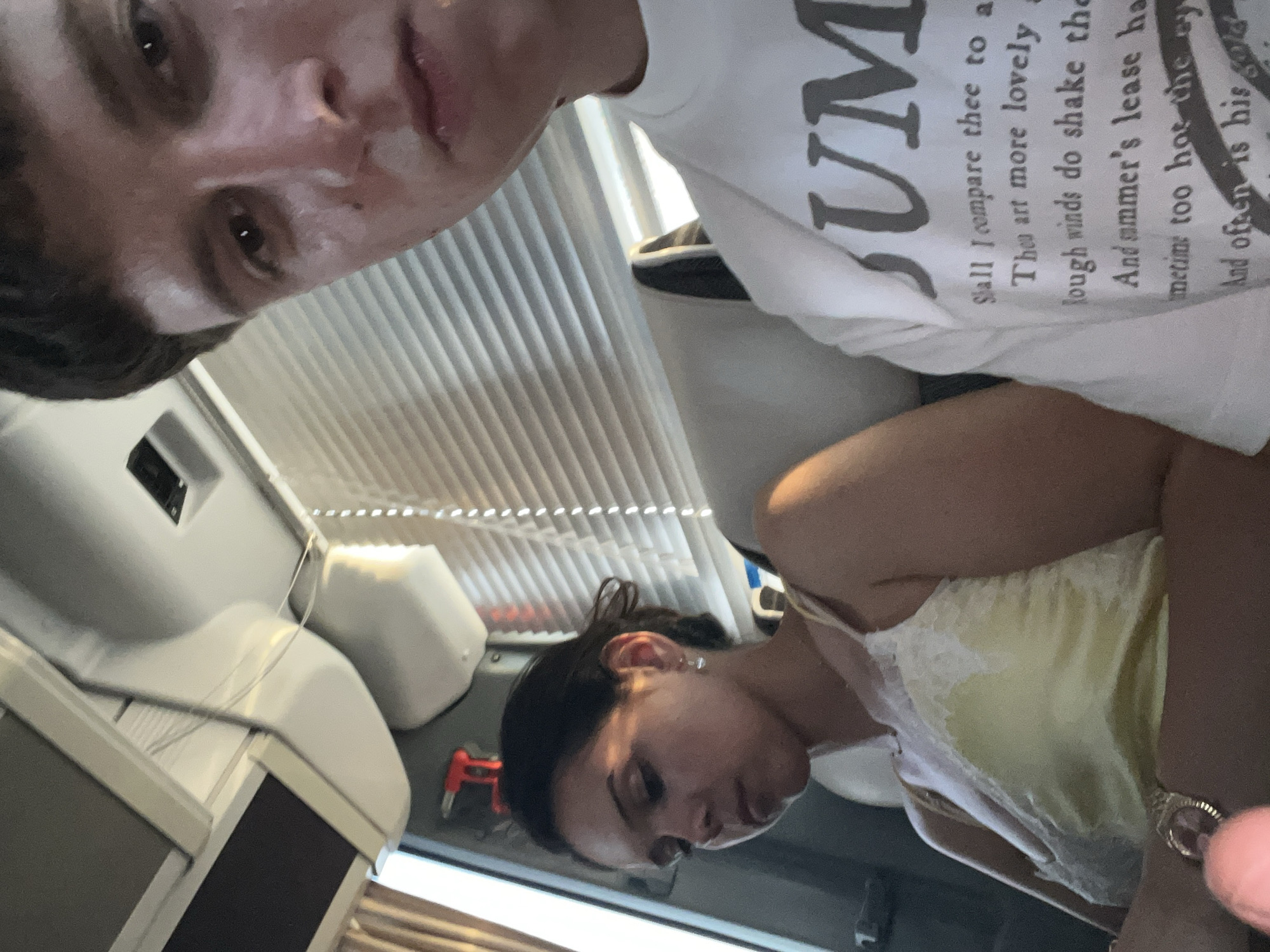 A selfie taken inside what appears to be a caravan or campervan. Two individuals are visible: A young man with short dark hair is at the top of the frame, angled down towards the camera. He wears a white t-shirt with printed text. Below him, a woman with dark hair pulled back is looking down. She wears a light yellow top with lace trim. Behind them are window blinds, a white molded interior wall, and a small red object attached to the wall. The location is consistent with the interior of a caravan that may be found at Ca' Savio, Italy.