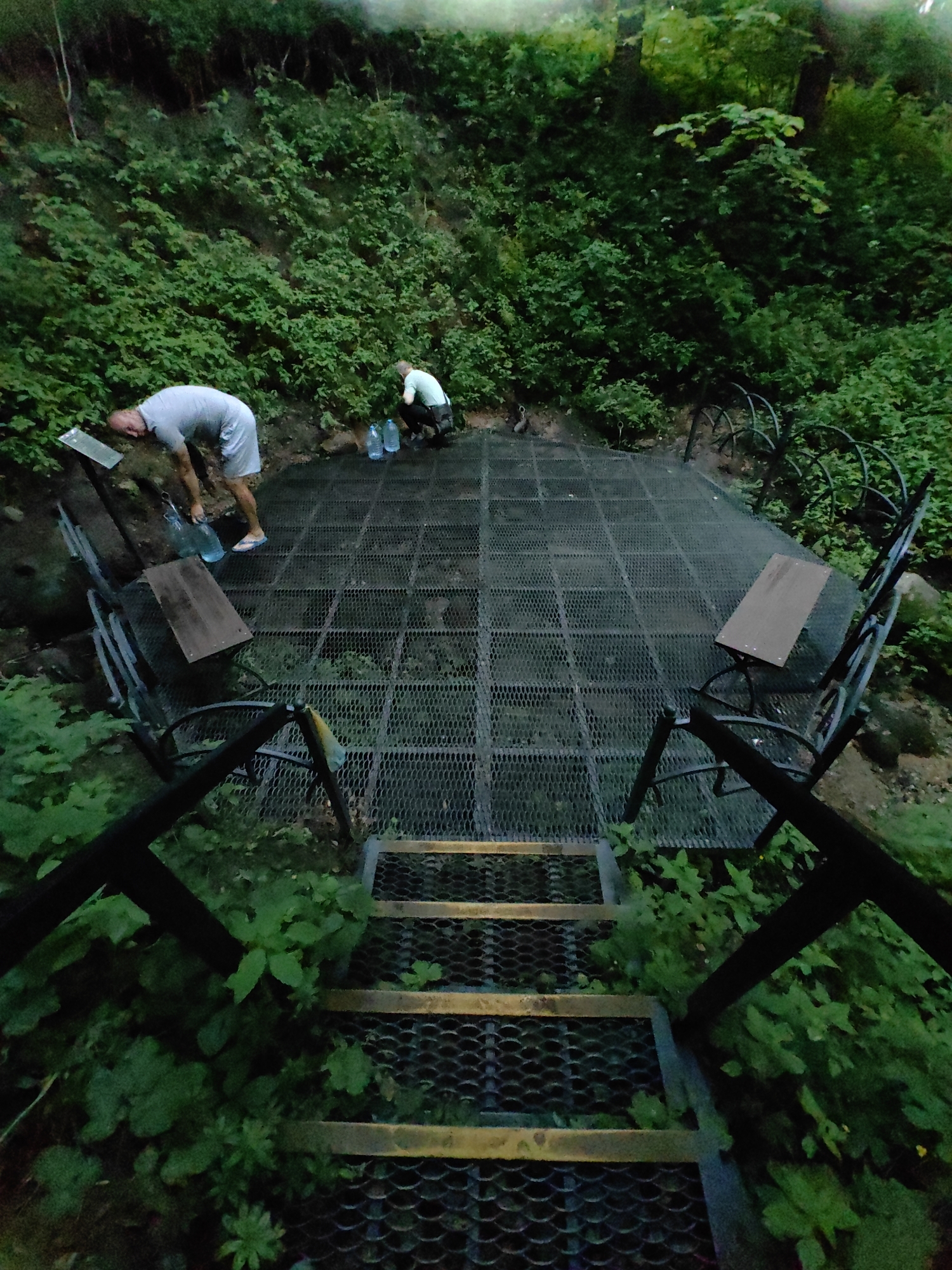 The image depicts an outdoor scene in a densely vegetated area, possibly a park or natural spring, located in Дзямідавіцкі сельскі Савет, Belarus. Two individuals are present. One person, wearing a grey t-shirt and shorts, is leaning over and appears to be filling or handling large clear plastic bottles. Another person, wearing a light-colored shirt and dark pants, is crouched down near some bottles.
The foreground features a metal grated staircase leading down into the scene. The main area is a metal grated platform with two benches. The platform is surrounded by lush green foliage and trees, indicating a natural setting. There are several clear plastic bottles on the platform, some of which appear to be filled with water. The lighting suggests it is either early morning or late evening, with a subdued, natural light.