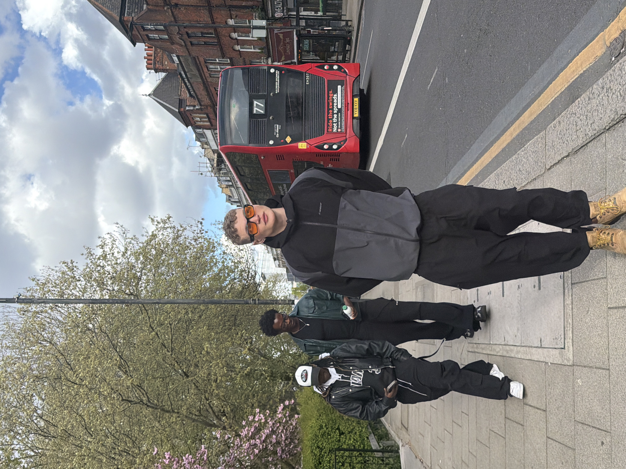 Three individuals stand on a sidewalk adjacent to a street in London, United Kingdom. A red double-decker bus, numbered "77," is visible in the background. Buildings flank the bus, with partial views of architectural details and signage. The sky is partially cloudy. A tree with new spring leaves and pink blossoms is positioned on the left of the frame.
The first individual, closest to the bus, wears a two-toned gray and black jacket, dark trousers, and tan boots, with orange sunglasses. Behind this person is a second individual in dark clothing. The third individual, positioned closest to the trees, wears a black and white jacket, dark trousers, and white sneakers, and has a white baseball cap. The subjects appear to be casually standing together in a group.
