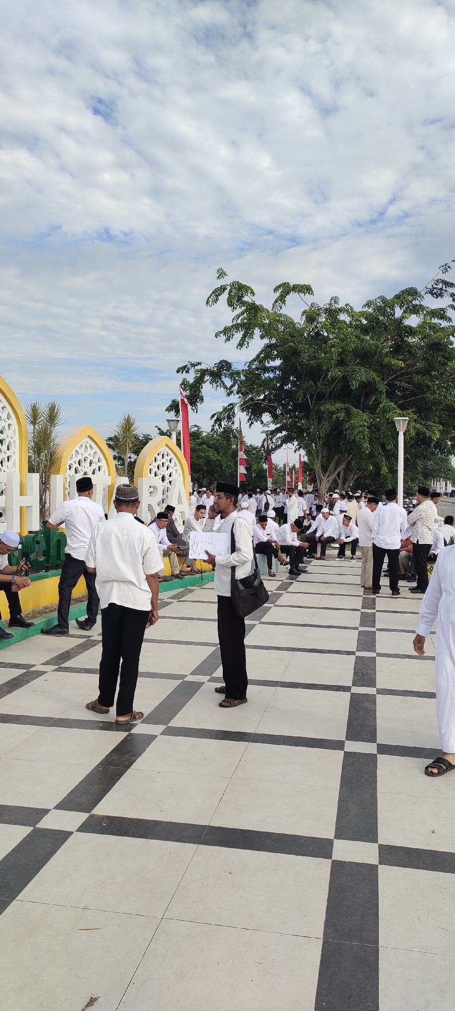 The image captures a gathering of people in what appears to be an outdoor public space, possibly in Meunasahaluedrienlhoksukon Barat, Indonesia. The ground is paved with a checkered pattern of light and dark tiles. In the background, there are large, decorative yellow structures with intricate white latticework, and several red and white flags are visible. A lush green tree dominates the upper right portion of the scene. A group of men, most wearing white shirts and many with traditional Islamic caps or headwear, are present. Some are standing, while others are seated on the ground or low structures. One man in the foreground is holding papers and speaking to another man. The sky is partly cloudy.