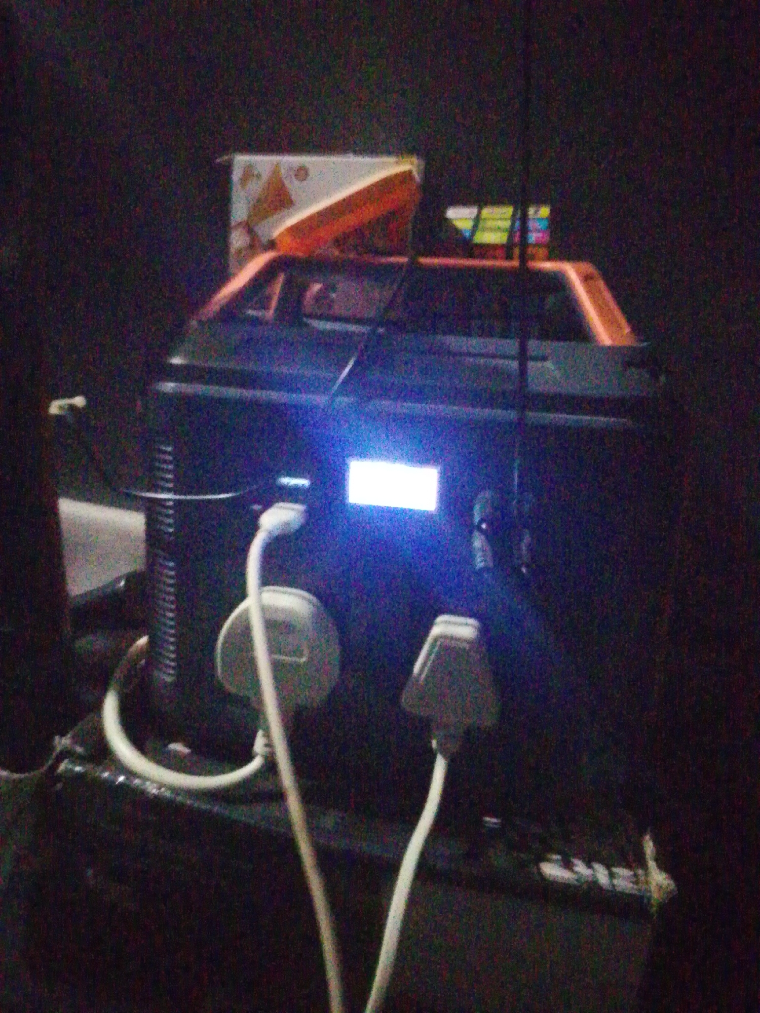The image displays a portable power station situated in a dimly lit indoor environment in the city of Unknown, Unknown. The device, positioned centrally, is dark-colored with a prominent orange top handle. Its front panel features an illuminated, unreadable rectangular digital display emitting a bright blue-white light, alongside a smaller, equally bright blue indicator light. Two white Type G electrical plugs are inserted into receptacles on the power station's front, with their cables extending downwards. The device rests on an indistinct, dark surface. In the upper background, a partially visible light-colored box with a triangular pattern is present, along with another object displaying horizontal multicolored stripes. The image quality is grainy and dark, indicative of low-light conditions. No individuals are depicted.