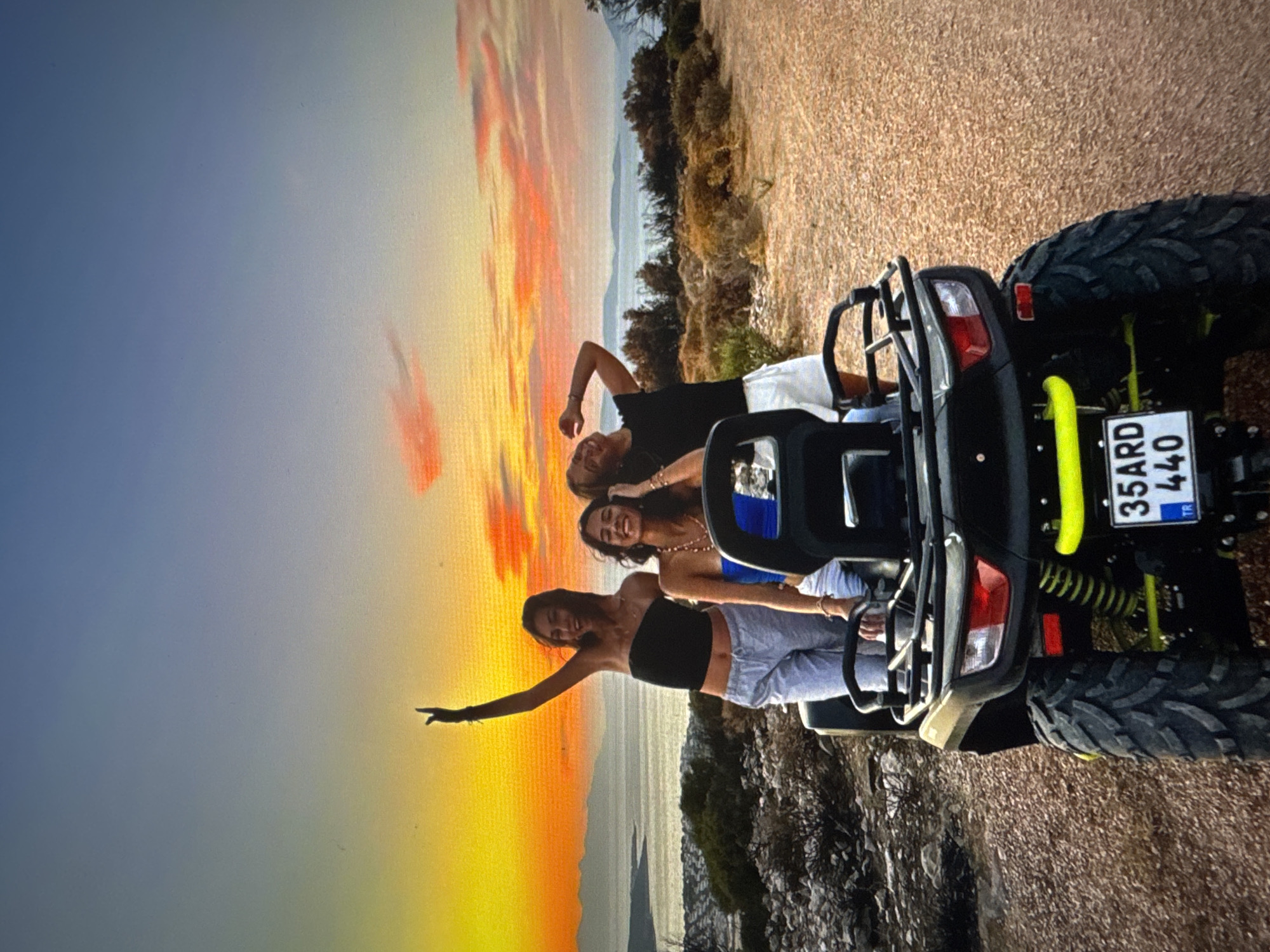 The image shows three young women on a quad bike at sunset. The woman on the left is wearing a black strapless top and light grey sweatpants, with her arm raised. The woman in the middle is wearing a blue top and white shorts. The woman on the right is wearing a black t-shirt and white shorts, with her head tilted back and hand on her head. They are on a dirt road with sparse vegetation. In the background, there is a body of water with islands and mountains, and the sky is a vibrant orange and pink. The quad bike is black with yellow accents and has a license plate that reads "35ARD 440 TR". The scene appears to be in a rural or coastal area. There is no indication that this location is in Tallinn, Estonia.