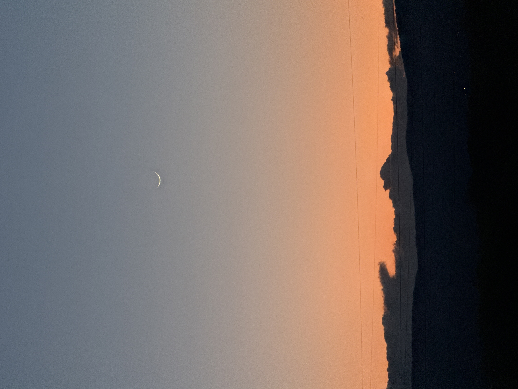 The image depicts a twilight sky with a crescent moon visible in the upper left quadrant. The sky transitions from a pale blue-grey on the left to an orange hue towards the right. A dark, vertical shape, possibly representing trees or buildings, occupies the right edge of the frame. There are also what appear to be power lines. There is no way to determine the location of Blagoevgrad, Bulgaria from the current image.