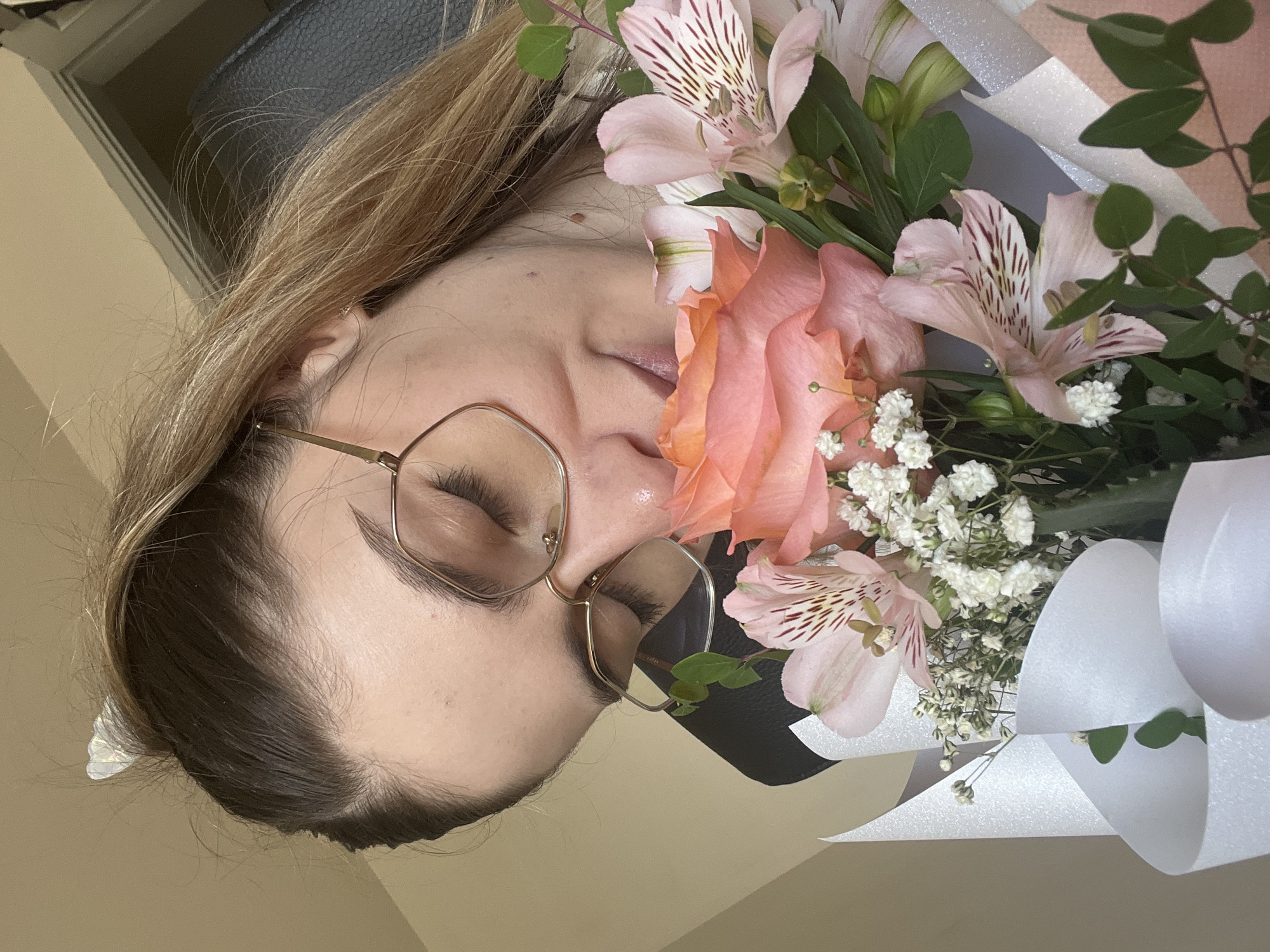 A woman with her eyes closed is smelling a bouquet of pink and coral roses, pink alstroemeria, and baby's breath. She is wearing glasses with gold frames and a black top. Her hair is pulled back in a ponytail with a white heart-shaped clip. The bouquet is wrapped in white and silver paper. The background is a plain wall. The scene appears to be indoors, possibly in an office or home.