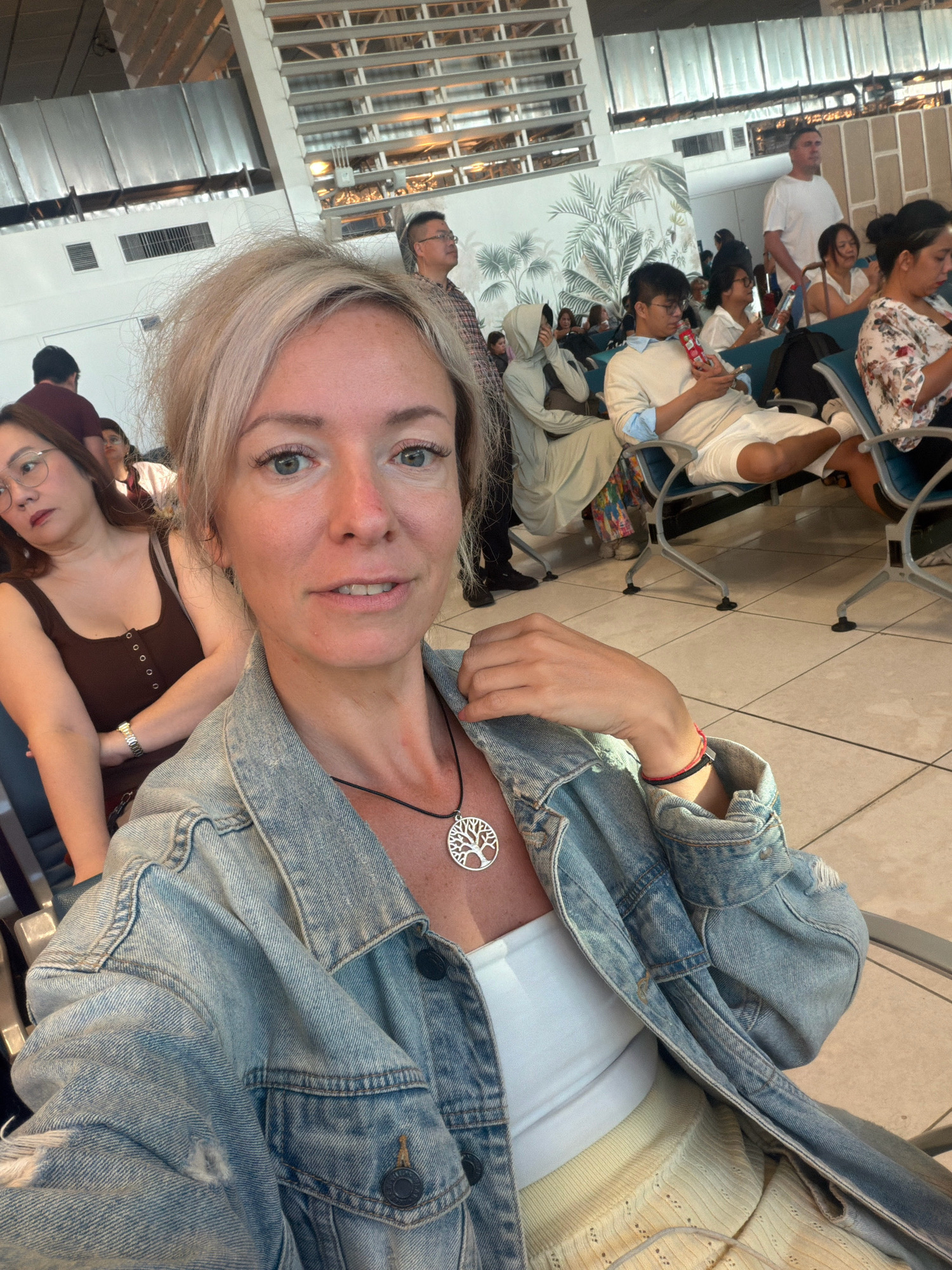The image shows a close-up of a woman with blonde hair, wearing a denim jacket and a white top. She has a tree-shaped pendant necklace. In the background, other people are sitting and standing in what appears to be an airport waiting area. The environment includes tiled flooring, rows of chairs, and a decorative wall mural with palm leaves. The location is identified as the Ninoy Aquino International Airport (NAIA) Terminal 3 in Pasay City, Philippines, suggested by the architectural style and the presence of travelers.