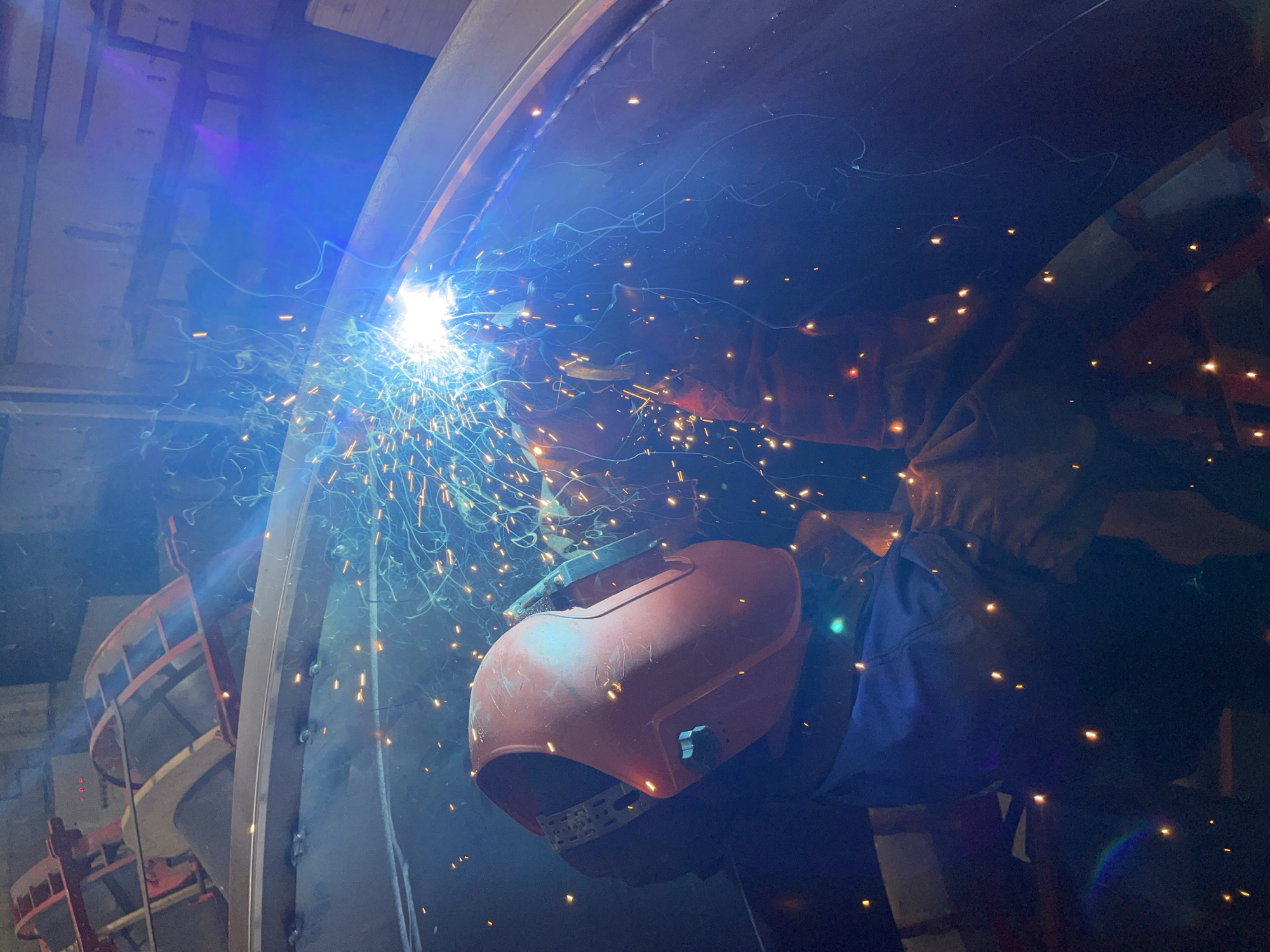 A welder, wearing a helmet and gloves, is welding a large metal structure. Sparks fly from the welding arc. The location is Čeluga, Montenegro. The welding appears to be part of a construction project. A red structure is visible in the background. The man is wearing a blue jumpsuit.