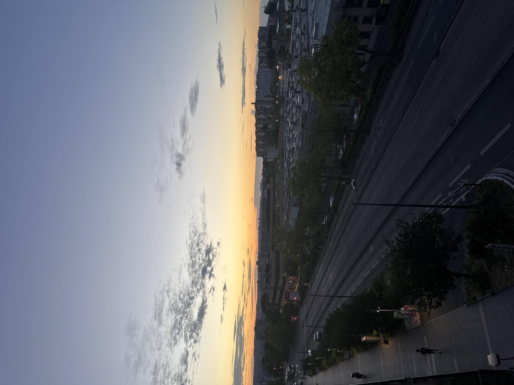 This is an aerial, high-angle shot of a city street at dusk. The sky is a gradient of pale blue at the top fading into a vibrant orange and yellow near the horizon, indicating the sun has recently set. Scattered clouds, tinged with grey and white, dot the upper portion of the sky.
Below the sky, a long, multi-lane road stretches into the distance, bordered by sidewalks and green spaces. The road is largely empty, with a few cars visible, and streetlights are illuminated. On the left side of the road, a sidewalk is populated by a single pedestrian running, and further down, a motorcycle is visible. Trees line the sidewalks and the median of the road, some with green foliage, others bare.
In the background, buildings of various heights and architectural styles are visible. Some appear to be residential apartment complexes with numerous windows, while others seem to be commercial or industrial structures with flat roofs and repeating patterns. The overall impression is a residential or mixed-use area within a city at the end of the day.