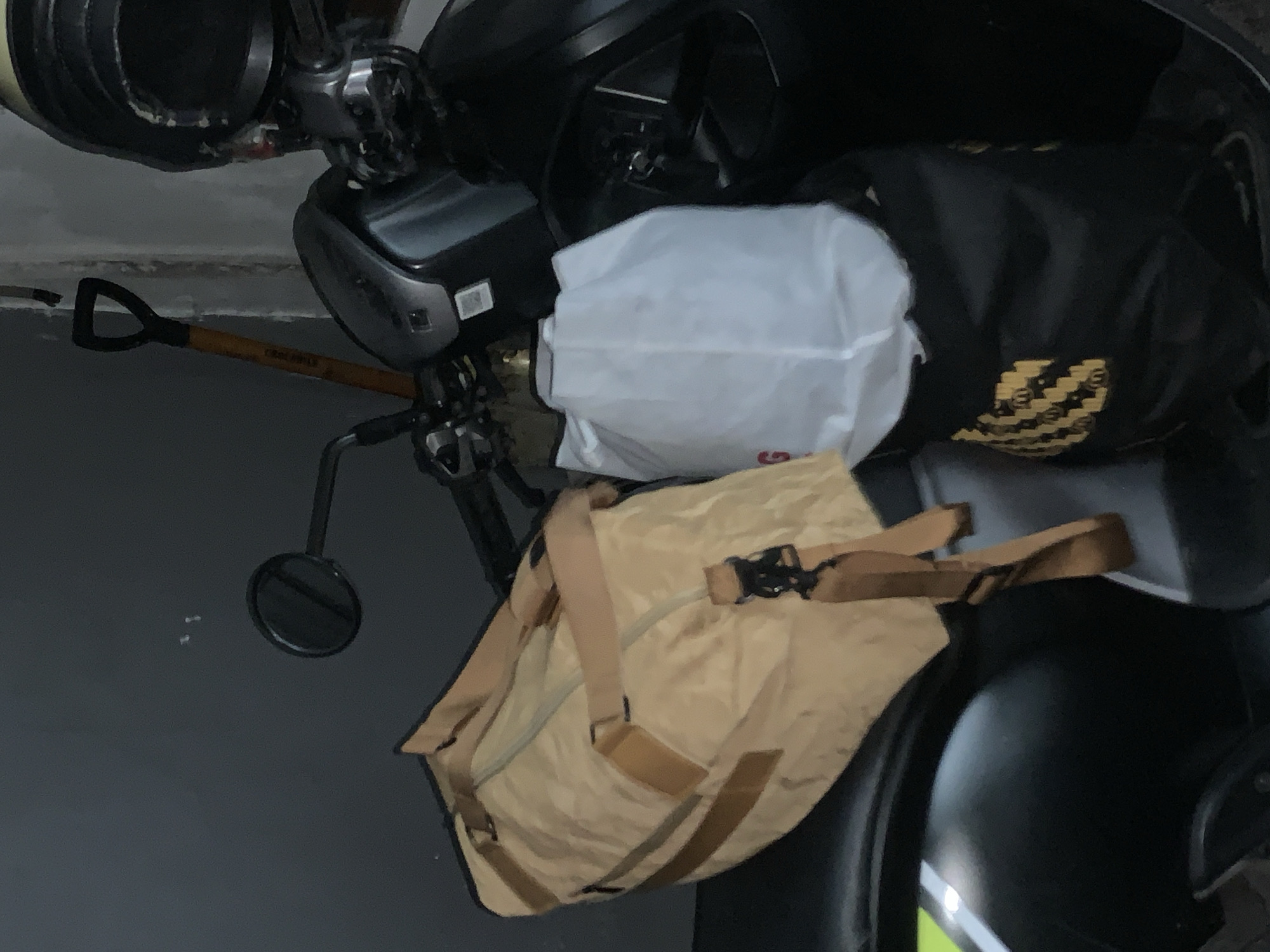 The image shows a close-up view of a black motorcycle parked outdoors, possibly in a garage or covered area, given the dark, muted background. The motorcycle is fitted with several items, suggesting it is being used for transportation or storage.
Prominently displayed on the motorcycle are two bags. One is a large black bag with a distinctive yellow and black patterned logo or design. To its left, partially obscuring it, is a white plastic bag, appearing to be a grocery bag or similar packaging. Below these bags and draped over the motorcycle is a tan-colored duffel bag. This bag has tan straps and a black buckle closure, indicating it's secured.
Other visible objects include a part of a shovel with a yellow handle and black grip, positioned horizontally across the lower left of the frame, leaning against the motorcycle. A round, black mirror, likely a motorcycle mirror, is also visible, attached to a stem.
The overall scene is dimly lit, suggesting it might be evening, early morning, or an indoor location with limited light. There are no people visible in the frame. The environment appears utilitarian, with a focus on the motorcycle and its cargo. The location context of Sukawati, Indonesia, does not provide specific visual cues within the image itself. No visible text other than the potential logo on the black bag.