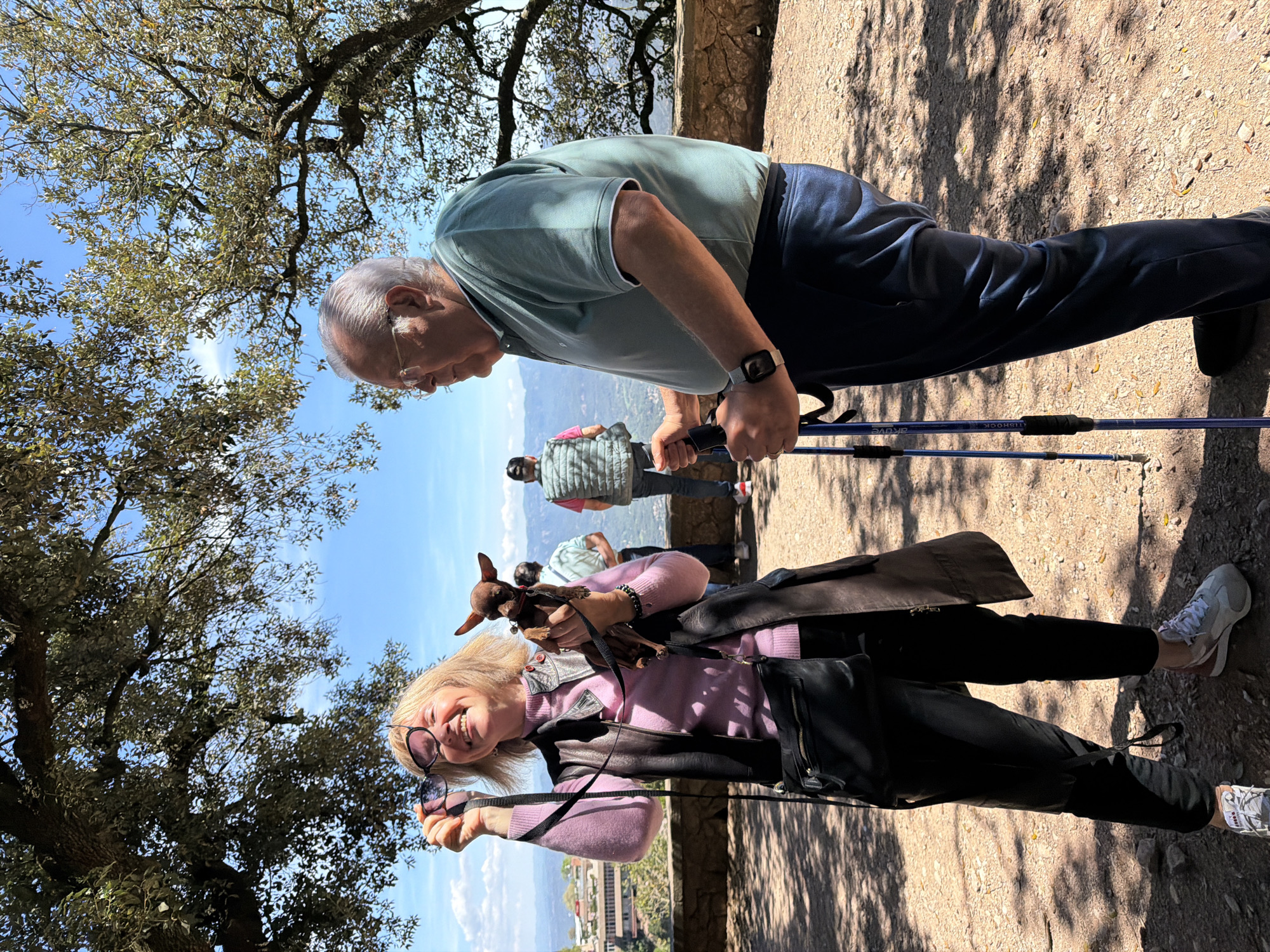The image captures an outdoor scene at a scenic mountain location, likely a path or viewpoint in Monistrol de Montserrat, Spain. The photograph is oriented vertically, depicting two main individuals and two others in the background under a bright, clear sky.
In the foreground, an older man with gray hair, wearing glasses, a light green polo shirt, and dark blue pants, leans forward, holding a pair of trekking poles. A watch is visible on his left wrist. He appears to be pausing or resting during a walk. Visible text on his trekking poles includes "AKUYE" and "ANTISHOCK".
Beside him, an older woman with blonde hair smiles brightly at the camera, holding a small brown dog, possibly a Chihuahua or Miniature Pinscher mix, in her arms. She wears a pink sweater, a dark jacket, and dark pants, with white and gray sneakers on her feet. She also holds a pair of dark sunglasses in one hand. A black shoulder bag hangs across her body.
In the background, two other individuals are partially visible further along the path; one wears a light green quilted vest. The ground is a light-colored dirt path with strong shadows cast by unseen objects, indicating bright sunshine and clear weather, likely midday or early afternoon. Overhead, the canopy of leafy green and brownish trees frames the blue sky. In the far distance, a mountainous landscape stretches under the bright sky, suggesting a panoramic view. The overall mood is one of enjoyment and relaxation during an outdoor activity.