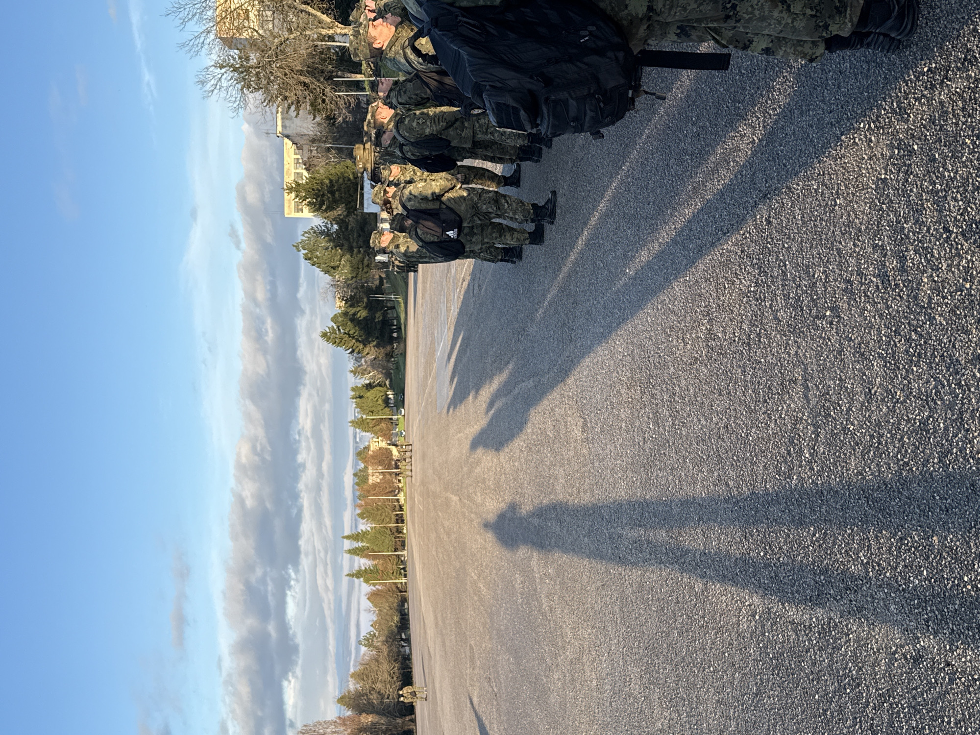 A line of people in camouflage uniforms stand on a gray, paved surface. Shadows extend from their positions. Beyond them, trees line the area, and a building is visible in the background. The sky is light blue with patches of clouds. The location is in an open area, likely a training ground or parade ground, in the city of Unknown, Unknown.