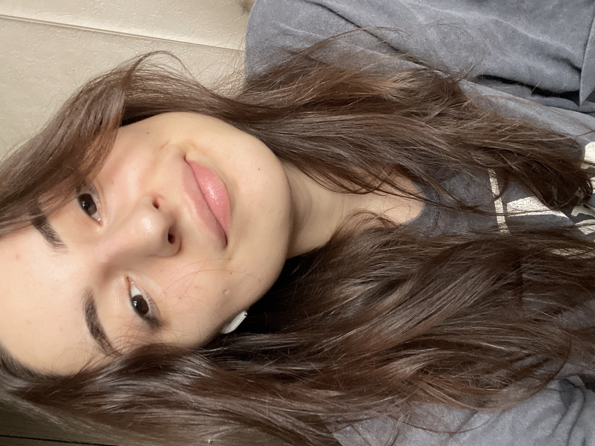 A close-up, slightly tilted selfie of a woman with long, brown hair. She is smiling slightly. She is wearing a gray shirt. One white earbud is visible in her ear. The background is out of focus, with a neutral-toned wall visible. No specific details suggest the location is Moscow, Russia.