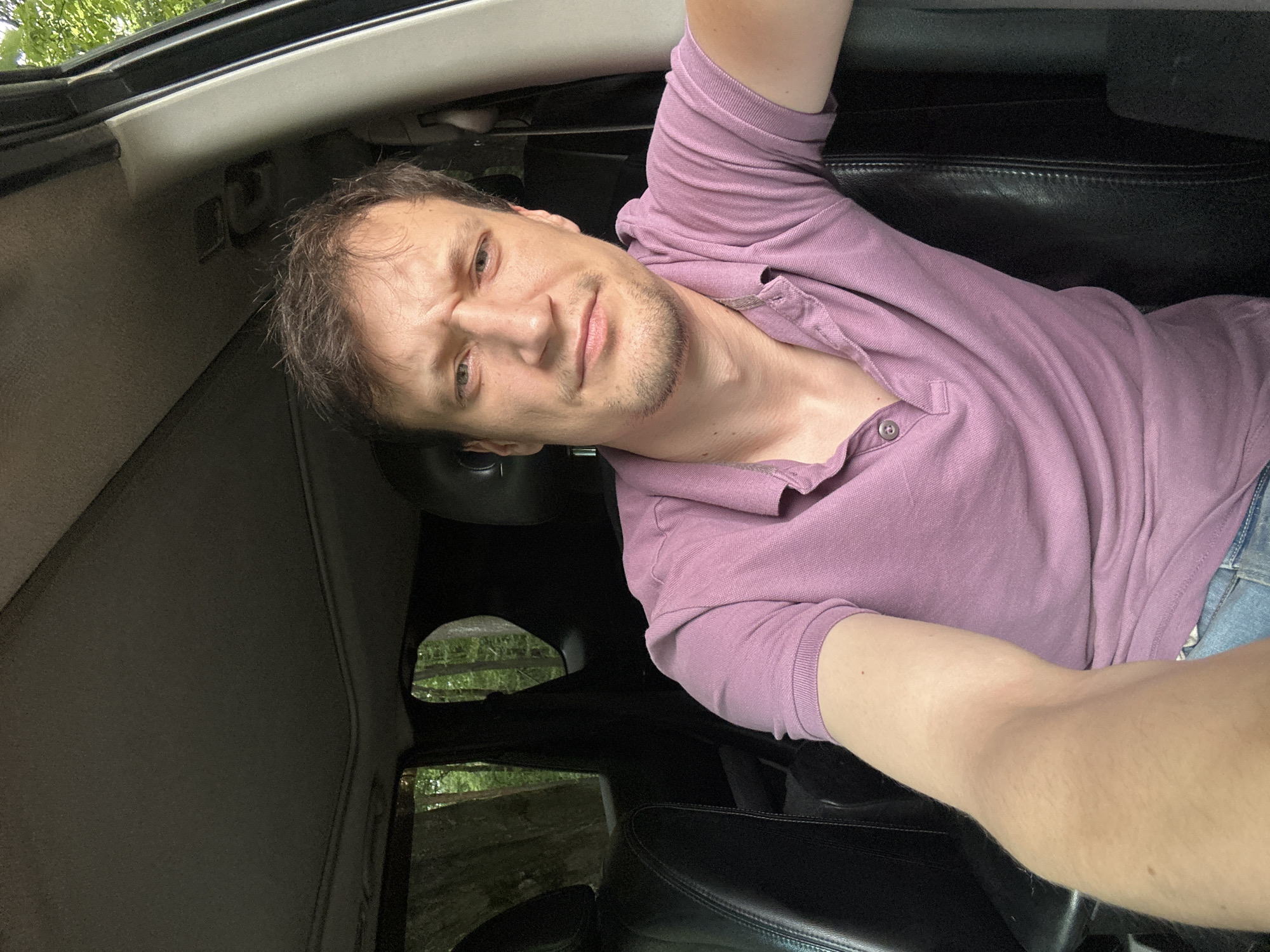 A man is sitting in the driver's seat of a vehicle, with his arm extended forward as if taking a selfie. He is wearing a purple polo shirt and has short brown hair and stubble. His expression is one of mild consternation. The interior of the vehicle appears to be dark leather. Through the windows, trees and foliage are visible, suggesting the vehicle is in a wooded area. The overall lighting suggests it is daytime. The location is not discernable from the image.