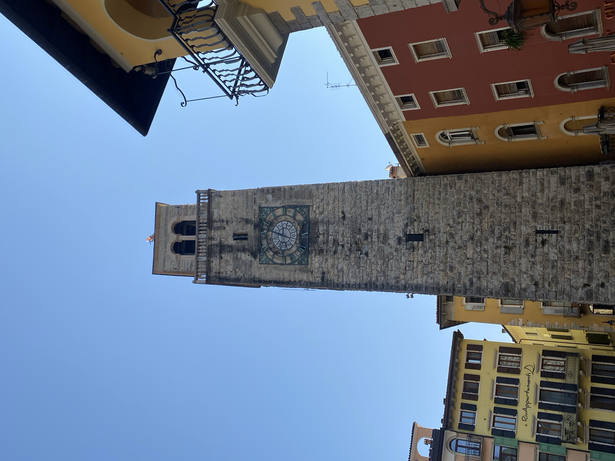 The image shows a view of the Torre Apponale, a medieval clock tower in Riva del Garda, Italy. The tower is made of stone and has a large clock face with Roman numerals. It is surrounded by colorful buildings with balconies and awnings. The sky is clear and blue. The scene appears to be a sunny day in a historic European town.