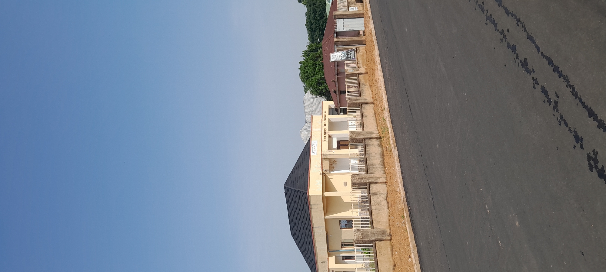 A wide shot shows a street in Jalingo, Nigeria, with a tan, single-story building to the left and a smaller, pink building to the right. The tan building has a dark gray, pitched roof and a white sign with black text that reads "RUTH PARTYED MULTI-PURPOSE HALL". White metal fencing runs along the front of the building, separating it from the street. The pink building to the right has a reddish-brown roof and a white sign with images and text. A paved road runs in the foreground, with tire tracks visible on its surface. The sky is clear and blue.