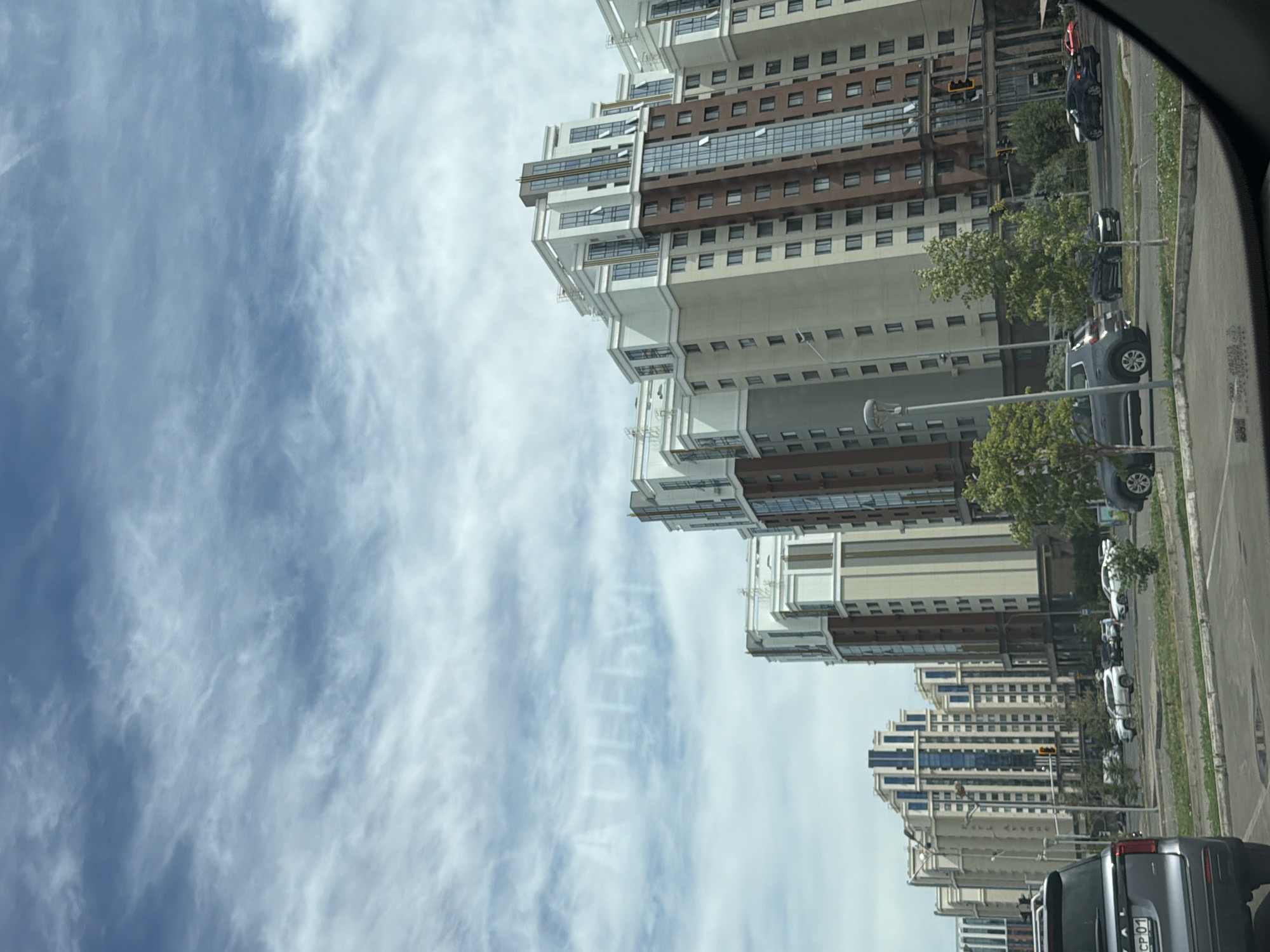 The image depicts a street scene in Astana, Kazakhstan, featuring a line of modern high-rise apartment buildings under a cloudy sky. The architecture of the buildings is characterized by their height, numerous windows, and a combination of beige, white, and brown facades, with some buildings featuring large glass sections. Several vehicles are visible on the road, including a dark gray SUV in the foreground. Trees and streetlights are also present, indicating an urban environment. The sky has faint writing in it, possibly from an airplane.