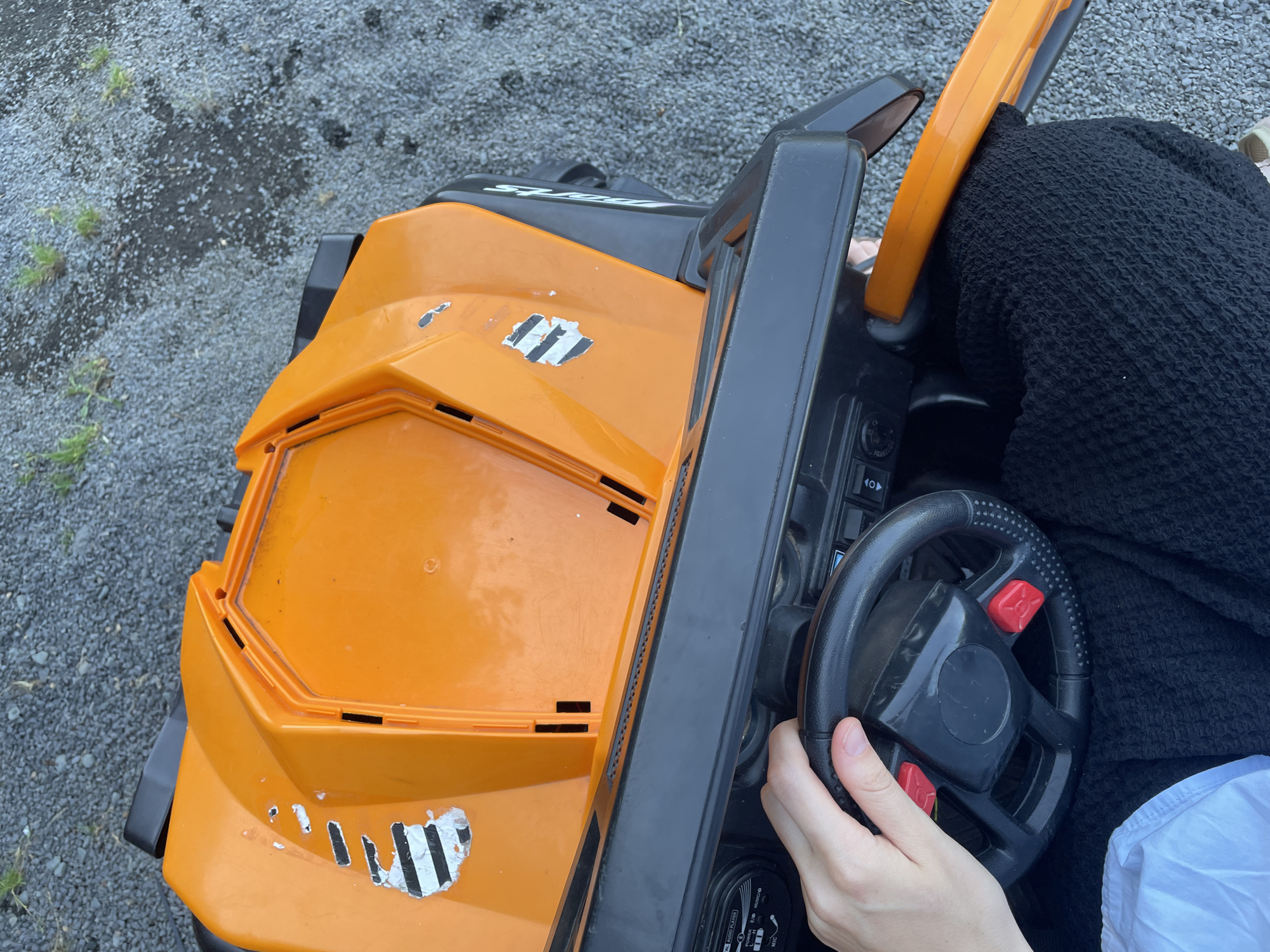An overhead, close-up shot captures a person's hand gripping the steering wheel of an orange toy vehicle. The vehicle is positioned on a gravel surface, likely a driveway or outdoor play area. The toy car's body is predominantly orange with black accents and features some wear and tear, including chipped paint and what appear to be remnants of racing stripes. A black dashboard with various buttons and a small screen is visible beneath the steering wheel. The person's hand is casually resting on the black steering wheel, with fingers slightly curled around it. Their attire consists of black textured fabric visible around the vehicle's interior, and a portion of a white garment is also visible. The background consists of small, gray gravel and sparse green vegetation. The location is identified as Beltershain, Germany.