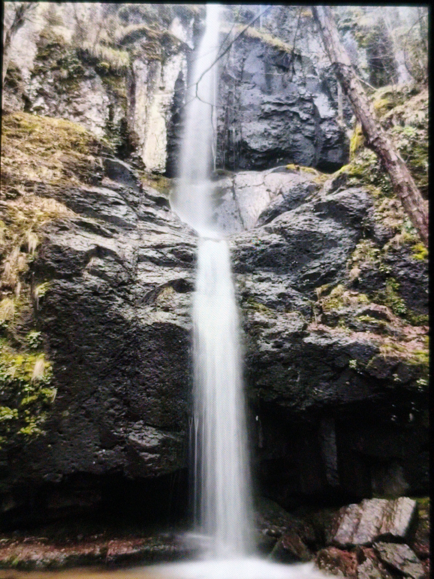 The image depicts a waterfall cascading down a rocky cliff face. The water appears smooth and blurred due to a long exposure. The rocks are dark and covered in moss and vegetation. The cliff face is surrounded by trees and foliage. The waterfall is located in the city of Devin, Bulgaria. There are no people visible in the image.
