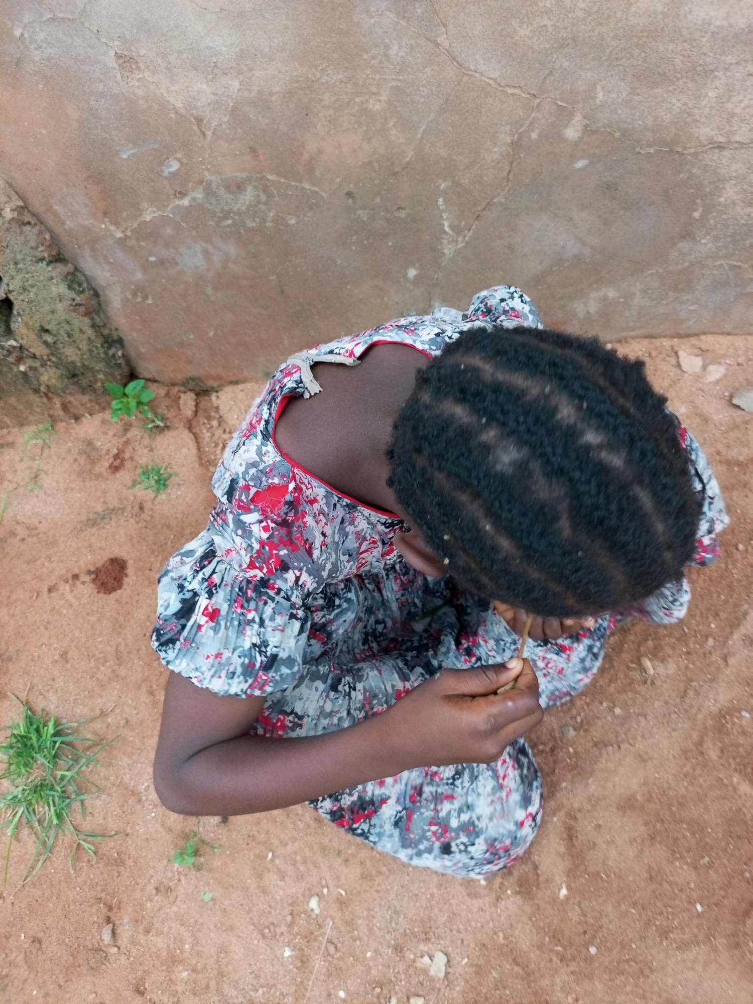 The image shows a young Black person from a high-angle perspective, sitting on reddish-brown dirt. The person is wearing a dress with a floral pattern of red, gray, and black. Their dark hair is styled in small braids or cornrows. They are holding a thin, twig-like object in their right hand. The background consists of a textured, light brown wall and some patches of green grass. The scene appears to be outdoors, possibly in a rural or less developed area. The lighting suggests it is daytime. This scene is set in Tempure, Nigeria.