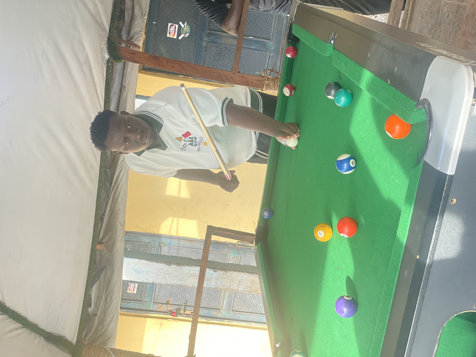 A young man plays billiards on a green billiard table in Lokoja, Nigeria. He wears a white t-shirt and stands in front of the table, cue stick in hand, preparing to strike the cue ball. Several colored billiard balls are scattered across the table. A building facade is visible in the background, featuring barred windows and an open door logo. The scene appears to be outdoors, under a tent or awning structure.