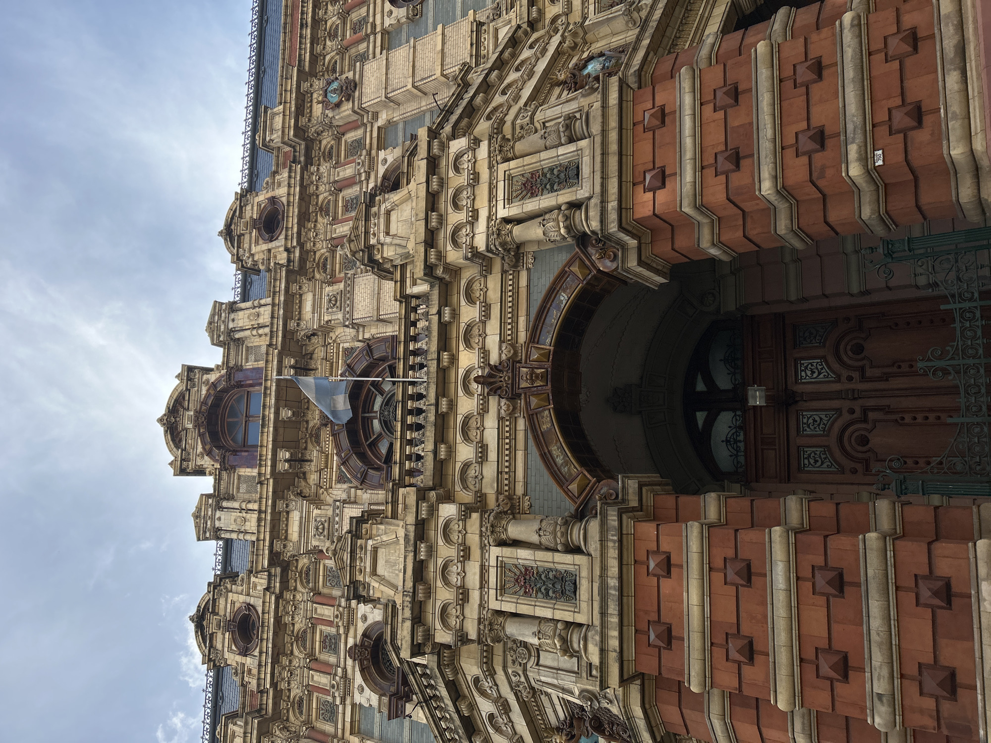 This image showcases the architecture of the Palacio de Aguas Corrientes, located in Buenos Aires, Argentina. The building's facade is visible, featuring ornate detailing, including brickwork, sculpted columns, decorative panels, and arched windows. A light blue and white flag is seen hanging from a flagpole. The sky is overcast, with light clouds.