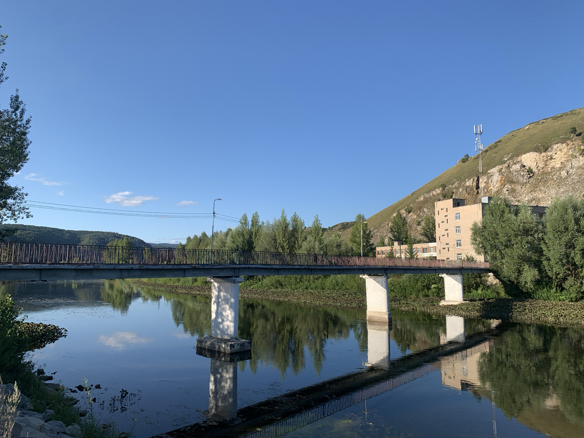 The image shows a bridge crossing a calm body of water, likely a river, in Asha, Russia. The bridge has a metal railing and is supported by two concrete pillars. The water reflects the bridge, trees, and a building situated on a grassy hill in the background. A communication tower is visible on top of the hill. The sky is clear and blue, with a few wispy clouds. The scene appears to be set during the daytime, with the sun casting shadows and illuminating the landscape.