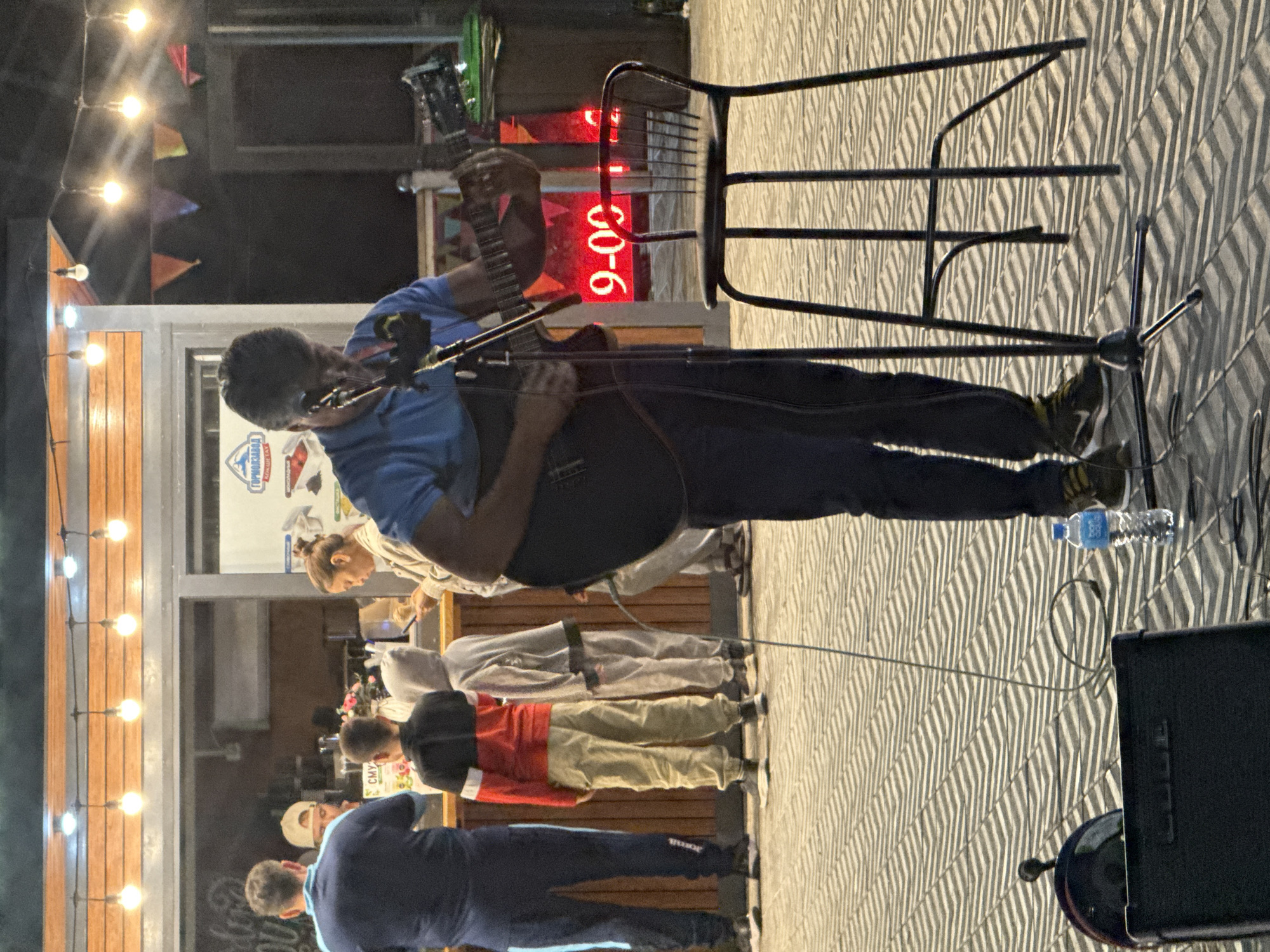 The image depicts an outdoor setting, likely a pedestrian area or a street with a patterned pavement. In the foreground, a man is playing an acoustic guitar and singing into a microphone, with a stool placed to his right. Behind him, a food or drink establishment is visible with people queuing or interacting. There are string lights and decorative flags overhead, suggesting it's an evening or night scene. A digital display behind the musician shows red numerals "9-00". The overall atmosphere suggests a public performance or event.