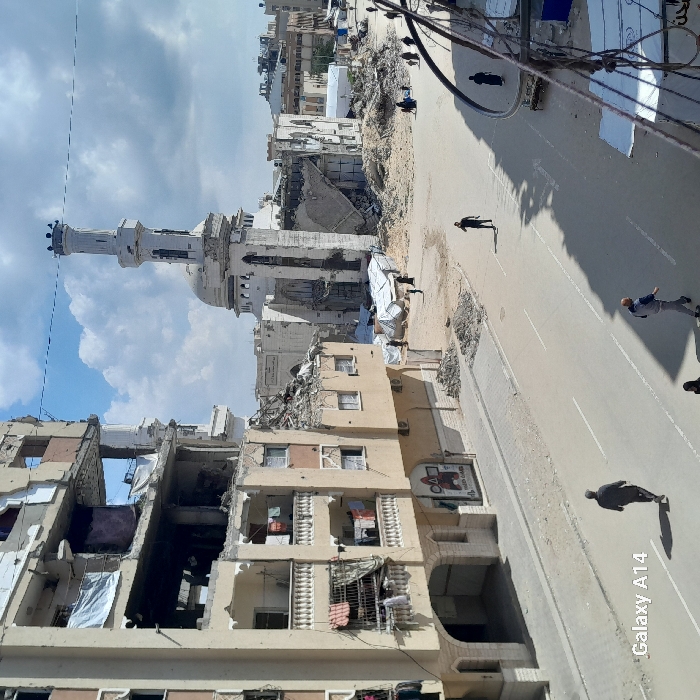 The image depicts a scene of extensive urban destruction in Khan Yunis, Palestinian Territory, under a partly cloudy sky. A multi-story building, heavily damaged with exposed interiors and missing sections, occupies the left side of the frame. Laundry or tarpaulins hang from several balconies of this structure, and a "SQUID GAME" poster is affixed to its lower level.
In the mid-ground, a wide, debris-strewn area extends towards a significantly damaged mosque, the focal point of the background. The mosque's minaret remains partially erect, though visibly damaged, while the main dome and surrounding structures show severe collapse and destruction. Scattered among the rubble near the mosque are several white tarpaulin-covered shelters.
A paved road with white lane markings runs through the foreground and mid-ground. Several individuals are visible on the road: one man walks away from the viewer on the left, another walks towards the right foreground, and several other figures are dispersed further down the street and near the makeshift shelters. Shadows are cast across the road and ground by the individuals and structures. Overhead wires are partially visible on the far right. The overall environment indicates widespread structural damage to buildings.