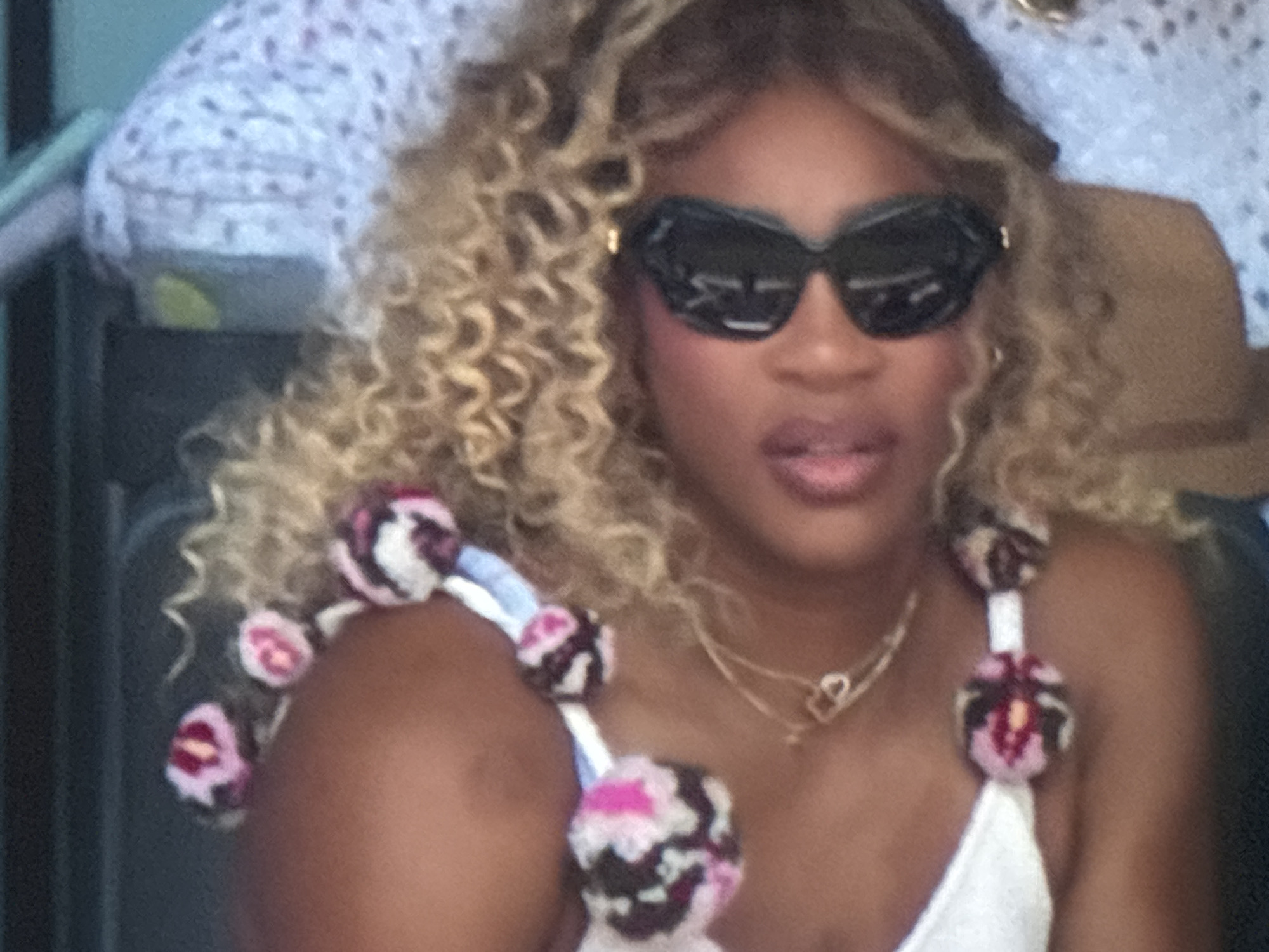 The image is a medium shot of a woman with blonde, curly hair and dark sunglasses. She is wearing a white top with decorative, pom-pom-like embellishments on the straps, which are pink, white, and black. A delicate gold necklace with a heart-shaped pendant is visible around her neck. The background is blurred but appears to be an indoor setting with a patterned fabric. No specific location in Burgas, Bulgaria can be identified from the image.