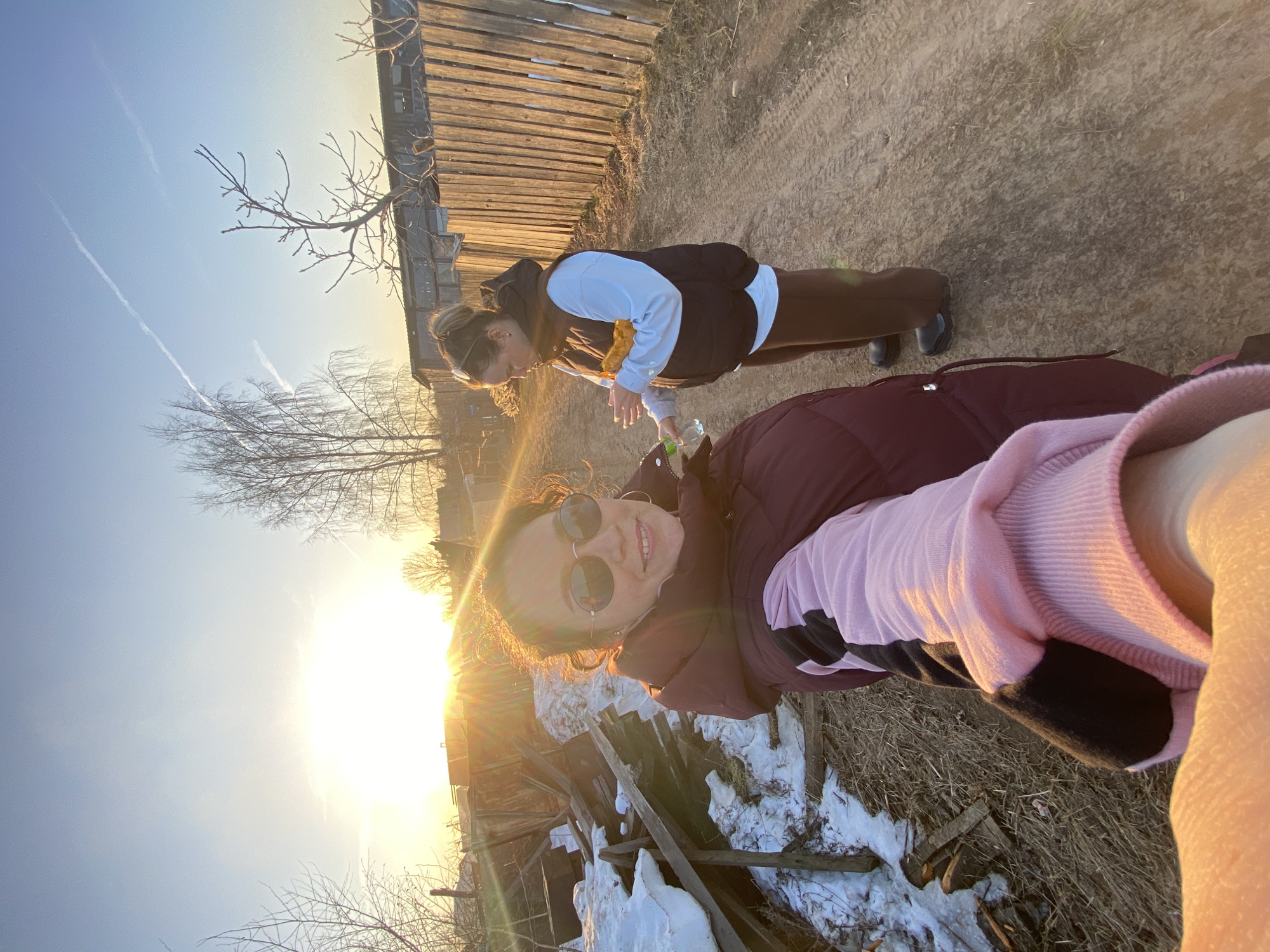 Two women are outdoors in what appears to be a rural or semi-rural environment. The woman in the foreground is holding the camera at a selfie angle. She is wearing sunglasses, a maroon jacket, and a pink sweater. The woman in the background is wearing a white long-sleeve shirt, a black vest, and brown leggings. Both women are fair-skinned. The setting sun creates a lens flare in the bottom left corner of the image. There is snow on the ground, as well as dry grass and debris. A wooden fence and trees are visible. The location is likely near Ольховка, Russia.