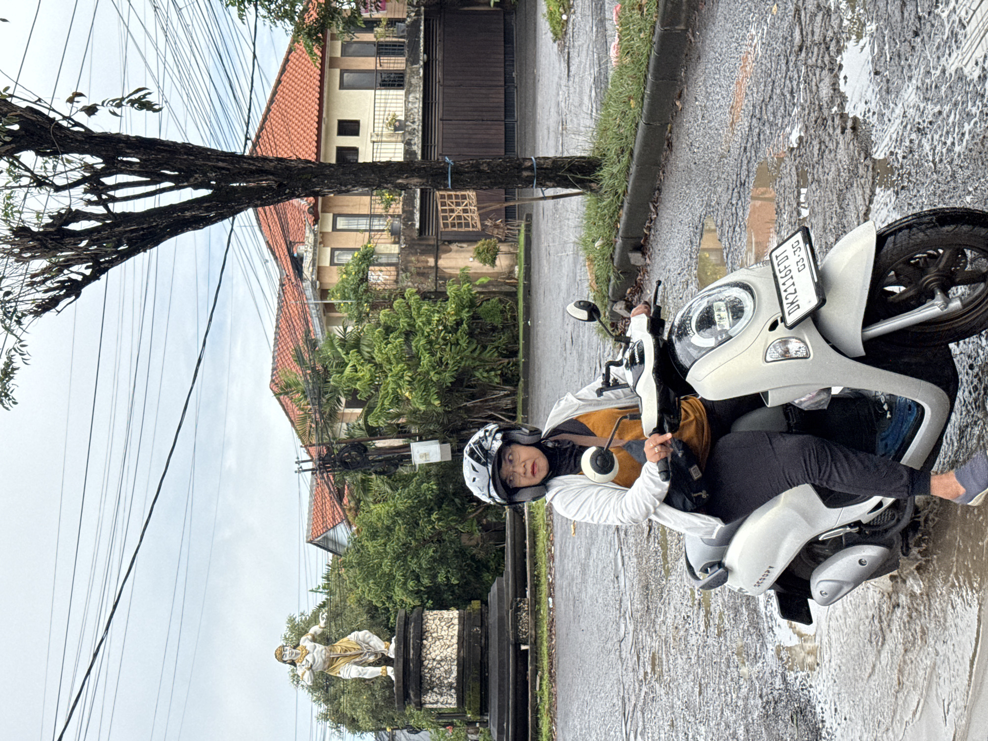 The image displays an outdoor street scene in Kuta Selatan, Indonesia, captured on an overcast day, likely after recent rainfall, as evidenced by the wet asphalt road and numerous puddles.
A person is the central subject, seated on a white Honda Scoopy scooter. They are wearing a white helmet with black patterns, a light-colored jacket over a brown/gold inner garment, and black pants. Their face is visible, looking towards the camera with a neutral expression. A brown backpack is slung over their shoulders. The scooter's front license plate clearly displays "DK 2116 FD" and "03-26." The word "SCOOPY" is also visible on the side of the scooter.
To the left of the person, a thick tree trunk rises, with its lower section tied with blue straps to a wooden support that holds a decorative, grid-like wooden cage. A network of black electrical or utility wires spans across the overcast, light grey sky. Further left and slightly behind the tree, a light-colored statue, resembling a Hindu deity or mythological figure, stands on a dark stone base amidst green foliage.
In the background, multi-story buildings with red tiled roofs and light-colored facades line the street, interspersed with lush green trees and bushes. A grassy curb runs along the edge of the wet road. The scene provides a glimpse into a residential or semi-urban environment in Indonesia during damp weather.