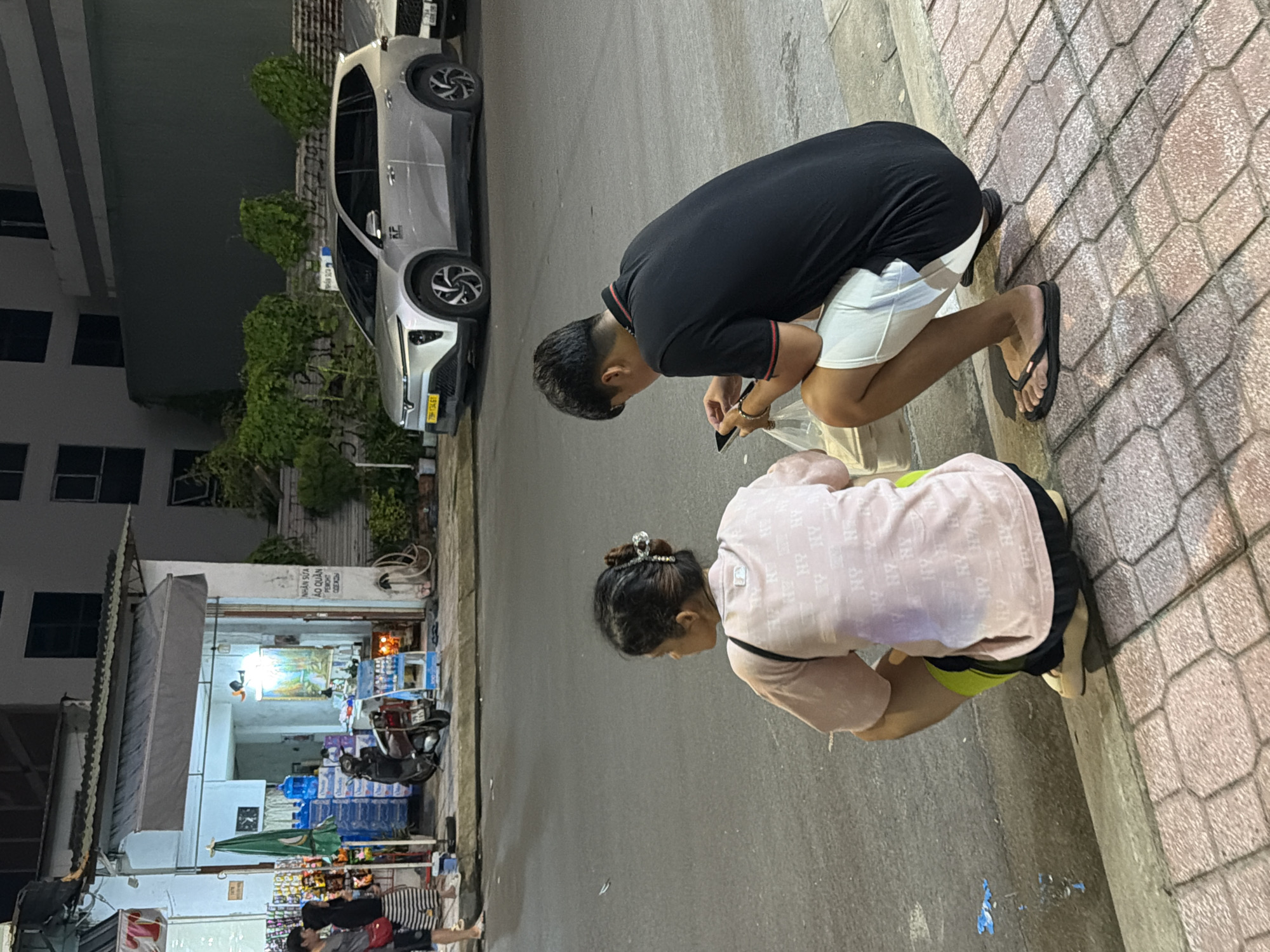 In Nha Trang, Vietnam, at street level, a woman with her hair in a bun is seen from behind, squatting with her knees bent and her hands on the ground. She is wearing a pale pink t-shirt with an all-over pattern of what appears to be "NY" letters and dark shorts. To her right, a man is also squatting and looking down at a smartphone in his hands. He is wearing a black polo shirt with red trim on the collar and sleeves, and white shorts. They are positioned on a sidewalk made of textured paving stones, next to a street. In the background, a small shop is visible, displaying various goods on shelves and in crates, including bottled water. A scooter is parked in front of the shop, and a silver SUV is parked further down the street. The lighting suggests it is evening or night.