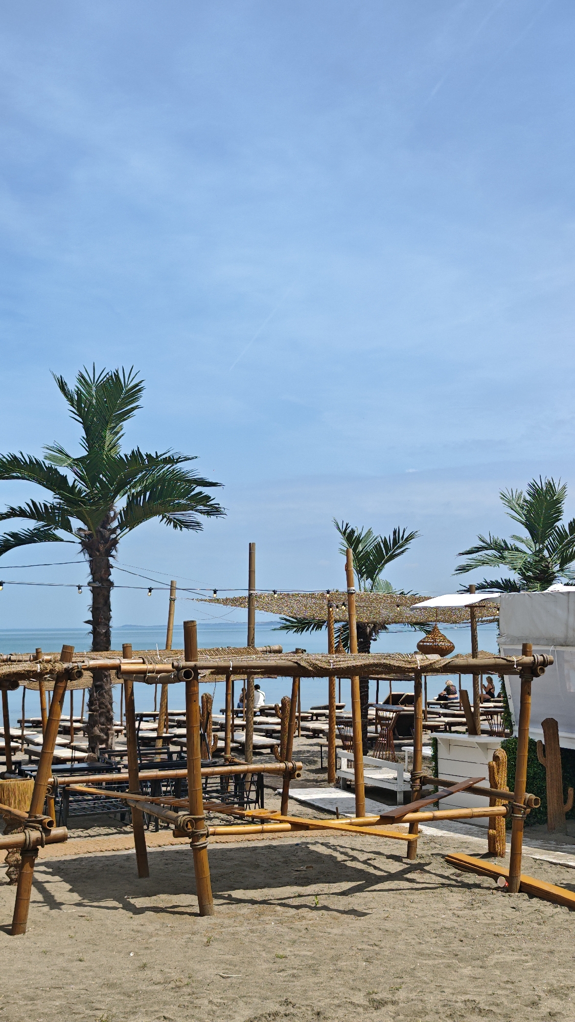 The image depicts a beach scene at the Budjaka Beach in Burgas, Bulgaria. The foreground features a bamboo structure, possibly a playground or seating area, built on the sand. Behind the structure, there are several rows of beach chairs and umbrellas, suggesting a beach club or resort. The Black Sea is visible in the background, meeting the clear blue sky. Three palm trees are also visible. Several people occupy the beach chairs.