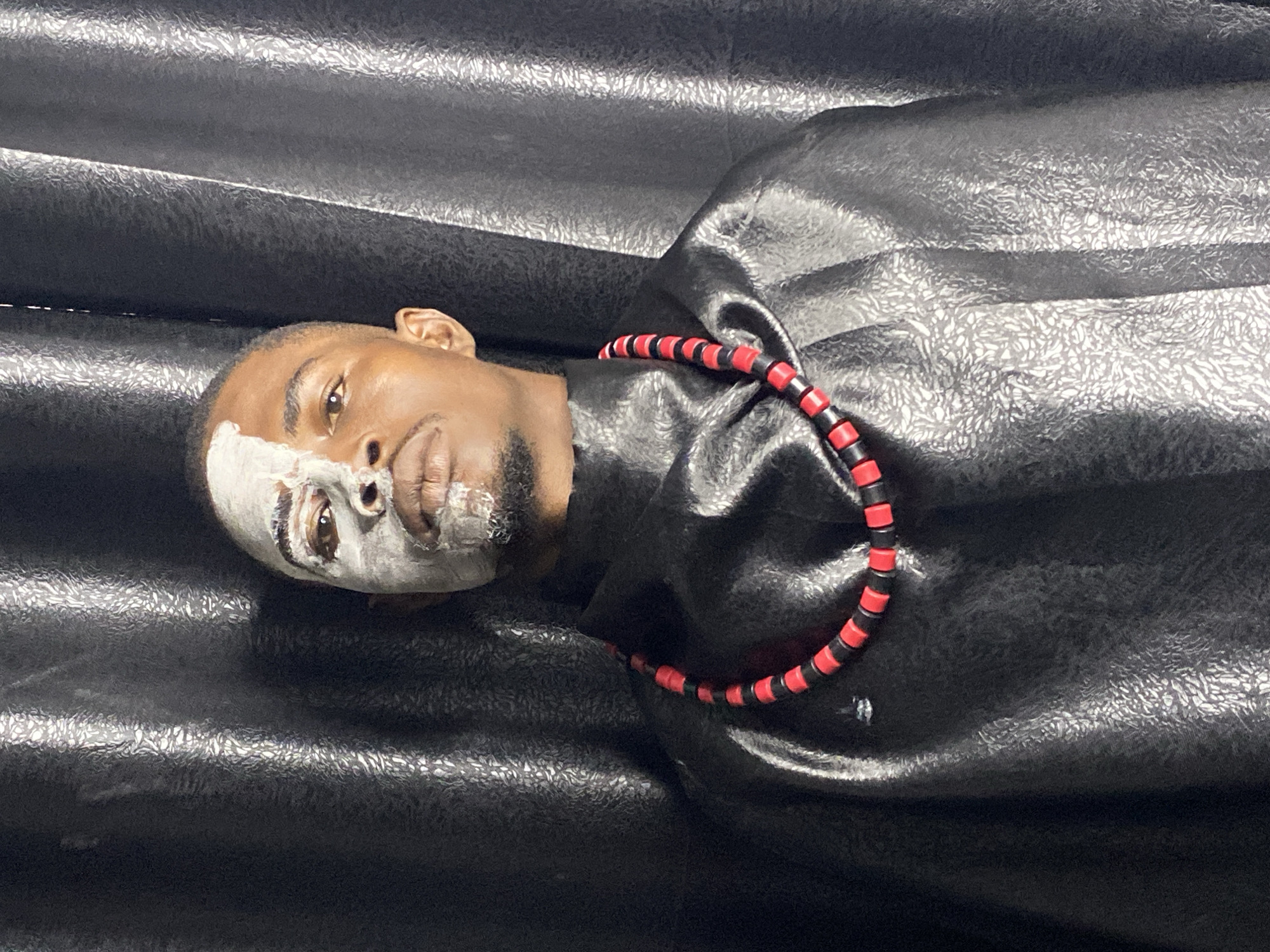 A portrait of a man with white face paint on the left side of his face stands against a dark, textured curtain. He is wearing a black garment and a necklace of red and black beads. The man is looking directly at the camera. The image was taken in Abakaliki, Nigeria.