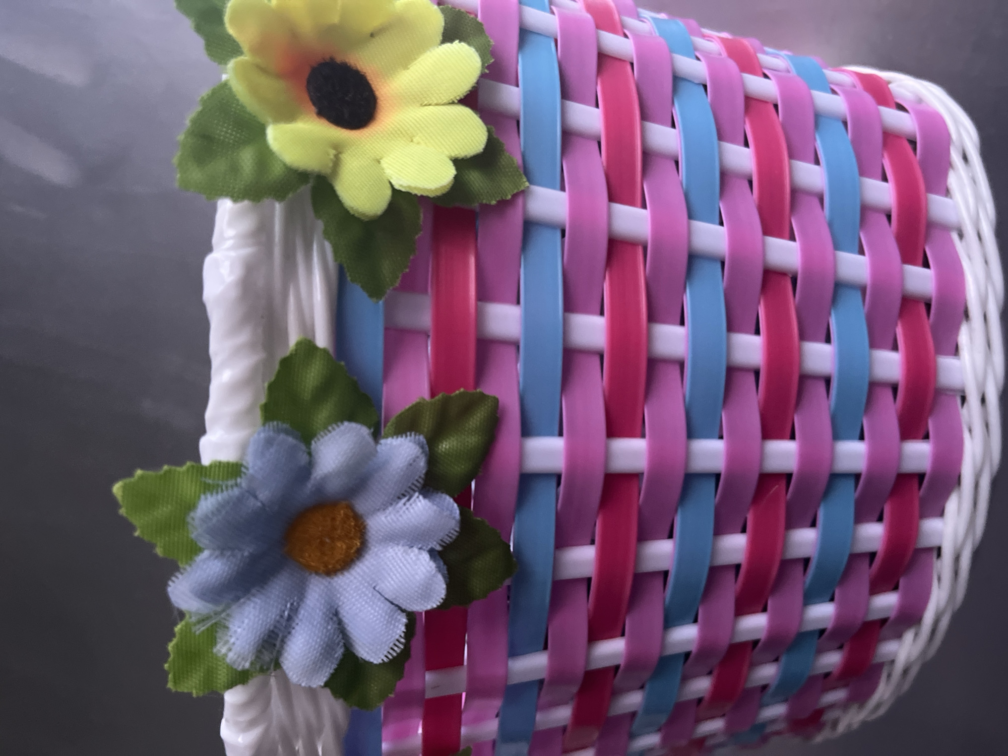The image shows a close-up of a small, decorative woven basket. The basket is made of interwoven strips of pink, blue, and white plastic. It is adorned with two artificial flowers: a light blue daisy with an orange center and green leaves on the left, and a yellow and orange daisy with a black center and green leaves on the right. The top edge of the basket is lined with white twisted material. The basket is resting on a flat, gray surface. The background is out of focus and appears to be a dark gray, possibly a wall or table. There is no indication of the basket's location in Garmisch-Partenkirchen, Germany, or any other specific place.