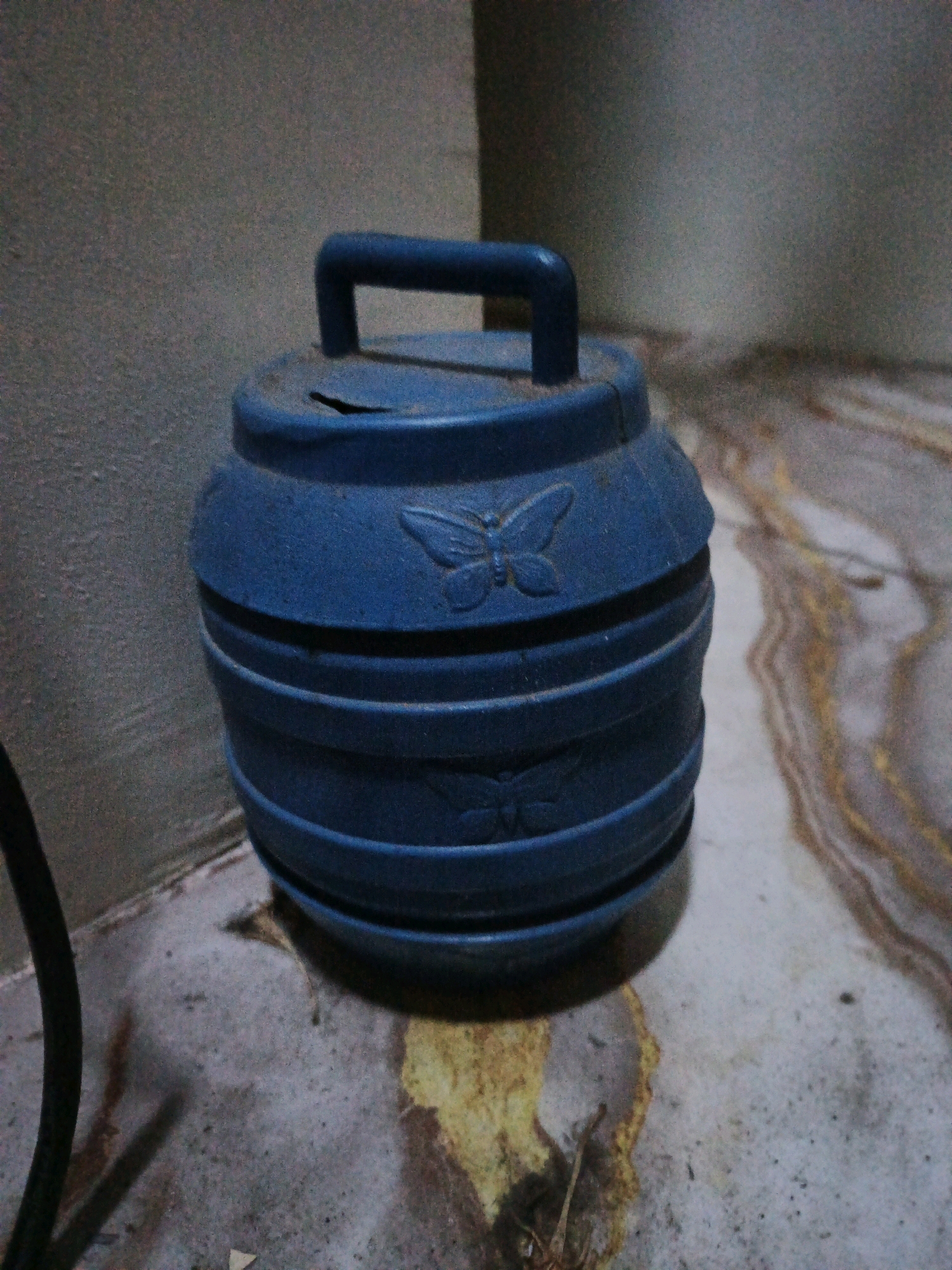 A blue, barrel-shaped piggy bank with a handle on top is placed on a dusty, tiled floor in a corner of a room. The piggy bank has horizontal ridges and two embossed butterfly designs. A dark cable is visible to the left of the piggy bank. The background consists of a textured wall. The scene is consistent with a residential setting in Ketegan, Indonesia, likely an indoor storage area or a less-trafficked corner of a home.