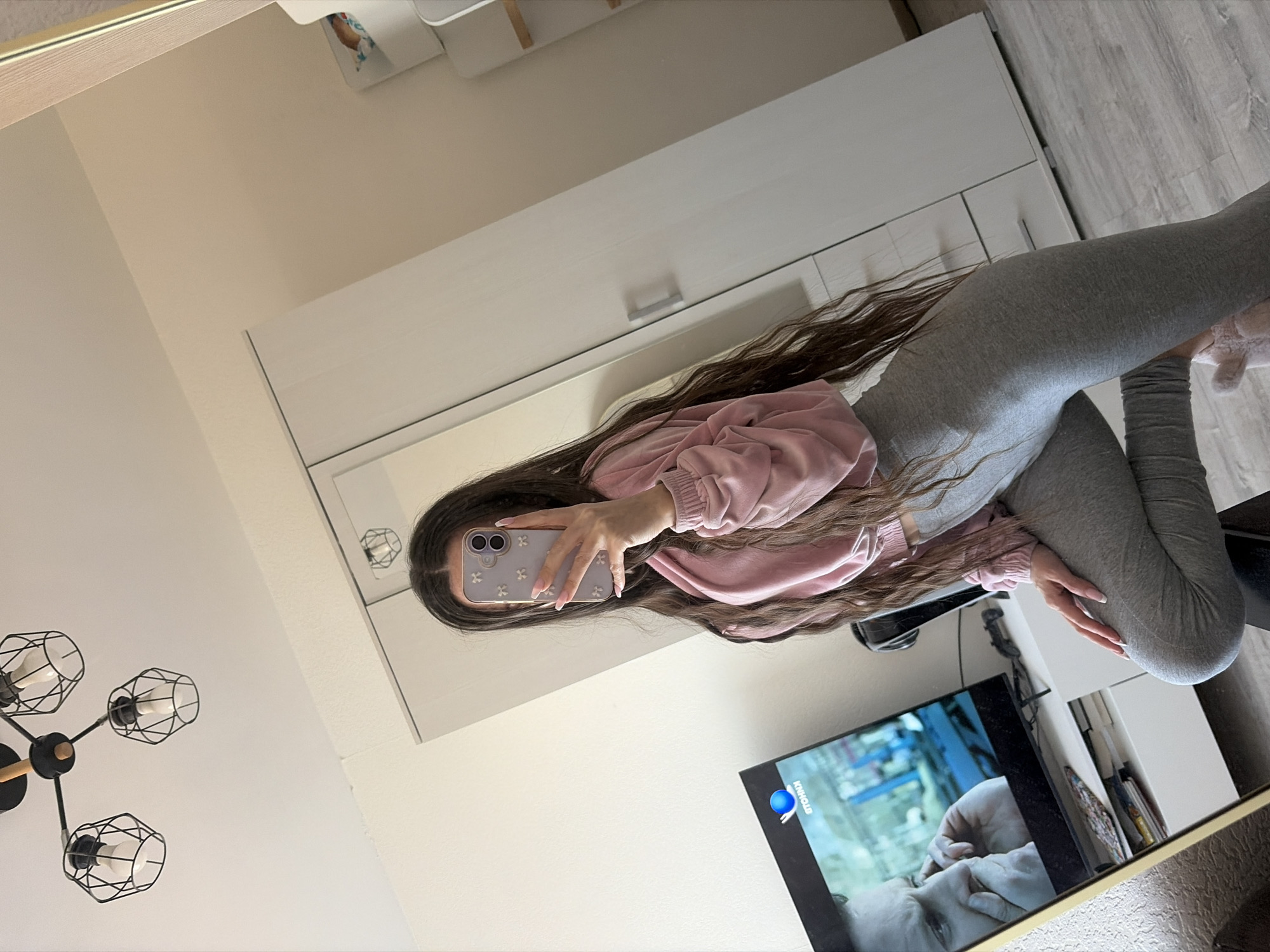 A young woman with long brown hair is seen in the reflection of a large mirror. She is wearing a pink sweatshirt and grey leggings. She is holding a smartphone with a light purple case, taking a selfie in front of the mirror.
The mirror reflects a portion of a room, which includes a television displaying a scene with a person's face, a modern chandelier-style light fixture, a white cabinet or shelving unit, and a glimpse of what appears to be a window or door. The reflection also shows a section of the floor, which has a wood-like pattern.
There is no indication of the location of this room being in Saint Petersburg, Russia.