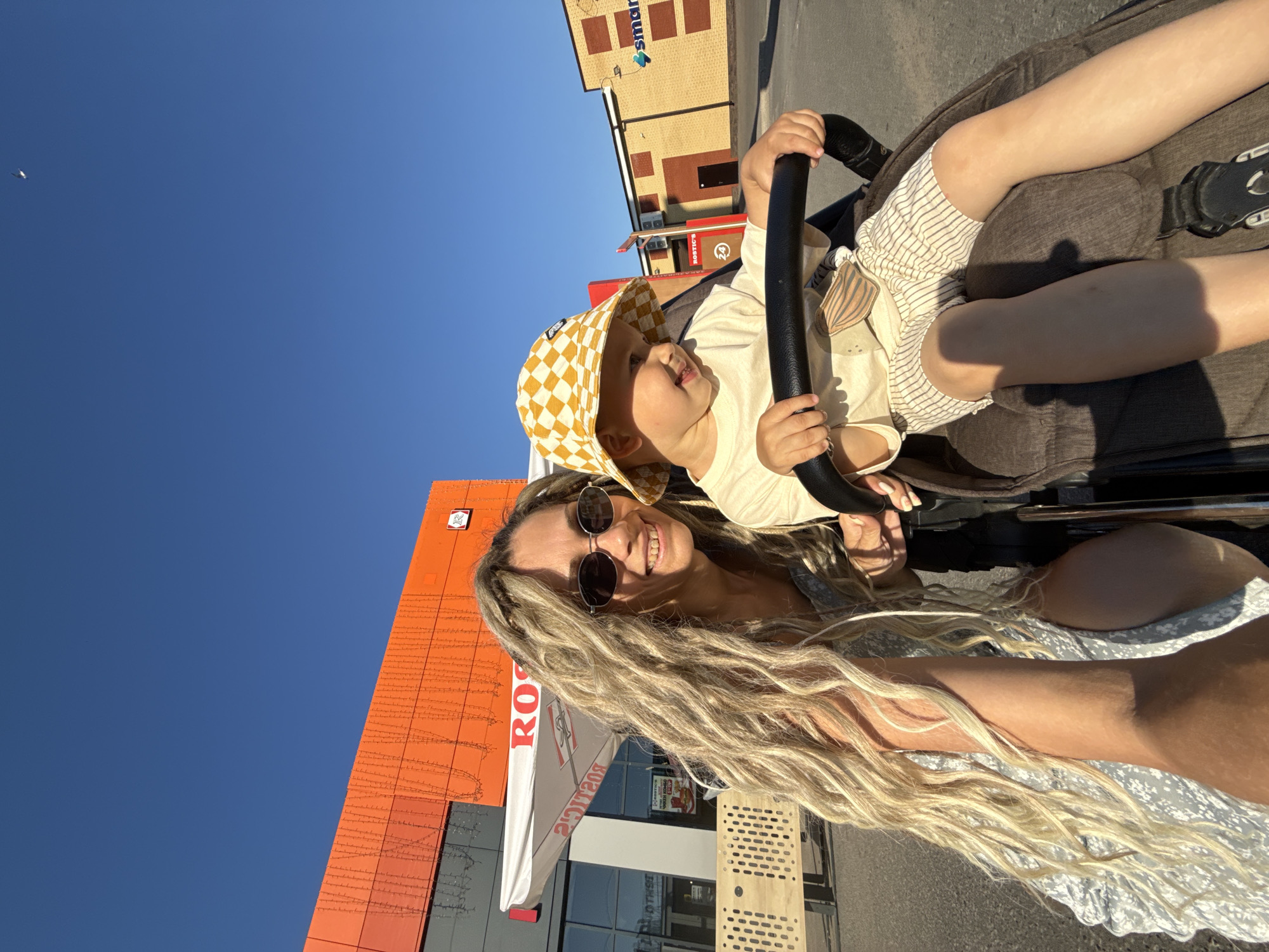 In this sun-drenched, outdoor scene, a woman with long, wavy blonde hair and sunglasses smiles as she sits beside a young child in a stroller. The child, wearing a yellow checkered bucket hat, looks to the side. They are positioned in front of a large, bright orange building with a white awning that has red lettering. In the background, a yellow brick building with a blue sign that reads "smart" is visible. The setting appears to be a commercial area, possibly a parking lot, under a clear blue sky. The overall impression is one of a casual outing on a warm day.