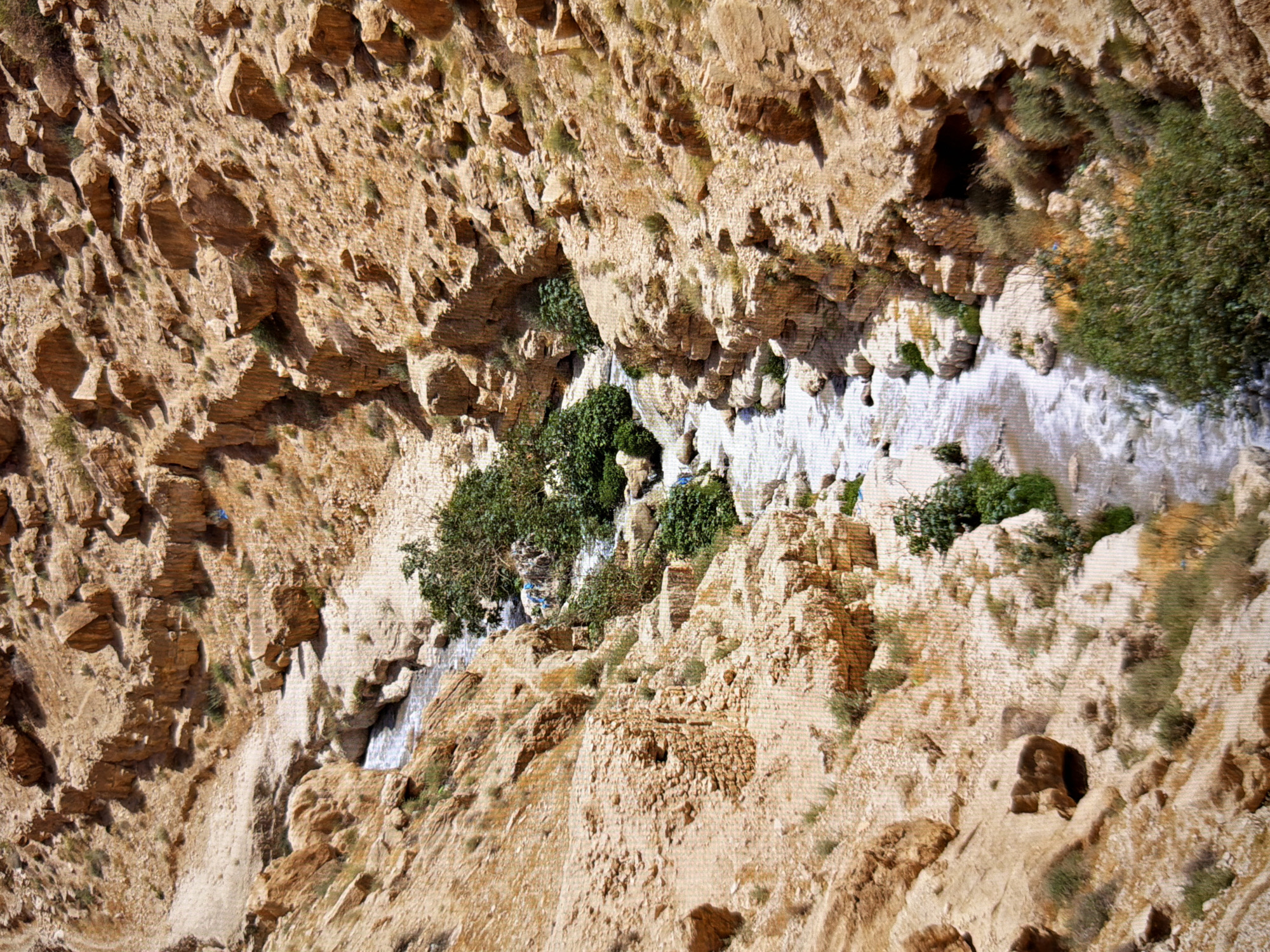 This image depicts a rugged, arid landscape with a narrow stream flowing through a canyon. The canyon walls are composed of layered, rocky sediment in shades of tan and brown, with sparse patches of dry vegetation and green shrubs clinging to the slopes. The stream appears shallow with visible rocks and some churning water, indicating a gentle flow. There are no people visible in the image, and no specific objects that can be identified beyond the natural environment. The scene conveys a sense of natural, untouched wilderness. There is no indication that this location is in Kyiv, Ukraine, or any other specific city.