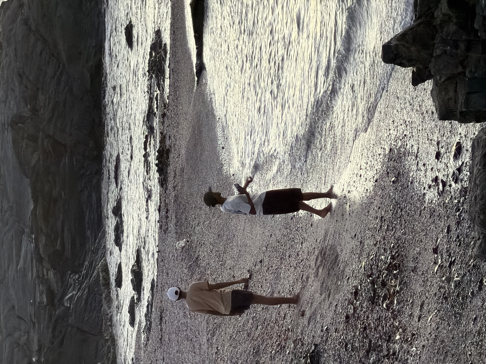 Two people are standing on a pebble-covered beach beside ocean water and under the face of a cliff. One individual is wearing a cap, T-shirt, and shorts, while the other person is dressed similarly. The location appears to be along a coastline with rocky features. Based on the topography and flora, the location could potentially be in Cape Town, South Africa, given the presence of cliffs and a coastal environment.