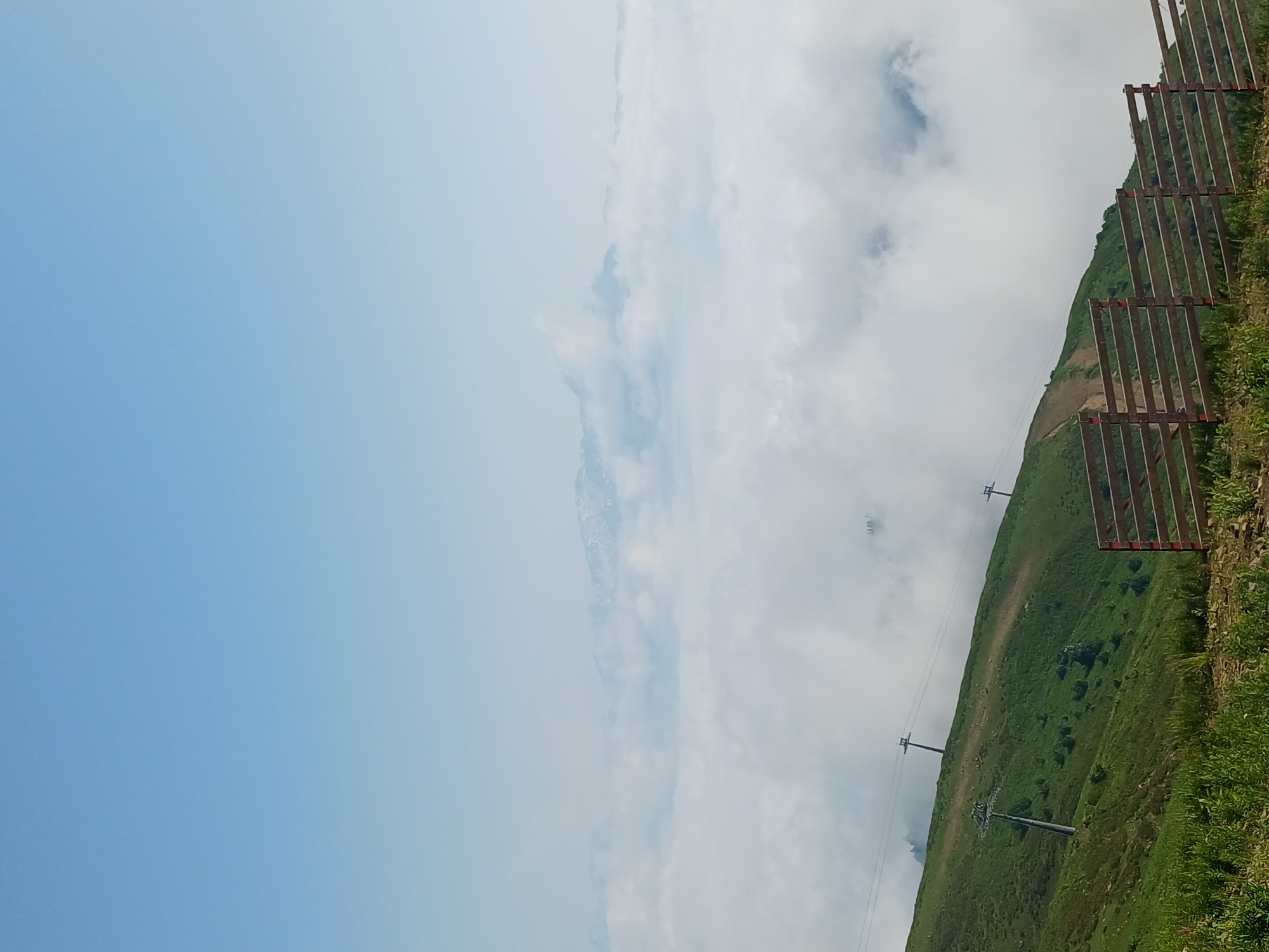 A photograph taken from a high elevation in Estosadok, Russia, showcases a mountainous landscape blanketed in thick white clouds. The sky above is a clear, pale blue. In the foreground, a lush green grassy slope descends towards the viewer. Several ski lift towers with cables are visible on the slope, indicating a ski resort or a similar recreational area. A wooden fence, constructed with horizontal planks and vertical supports, runs along the lower right portion of the frame, partially obscuring the lower part of the slope. In the distance, the jagged peaks of snow-capped mountains emerge from the sea of clouds, hinting at the vastness of the surrounding mountain range. The overall scene conveys a sense of natural beauty and elevation, with the clouds creating a dramatic visual separation between the foreground and the distant mountains.